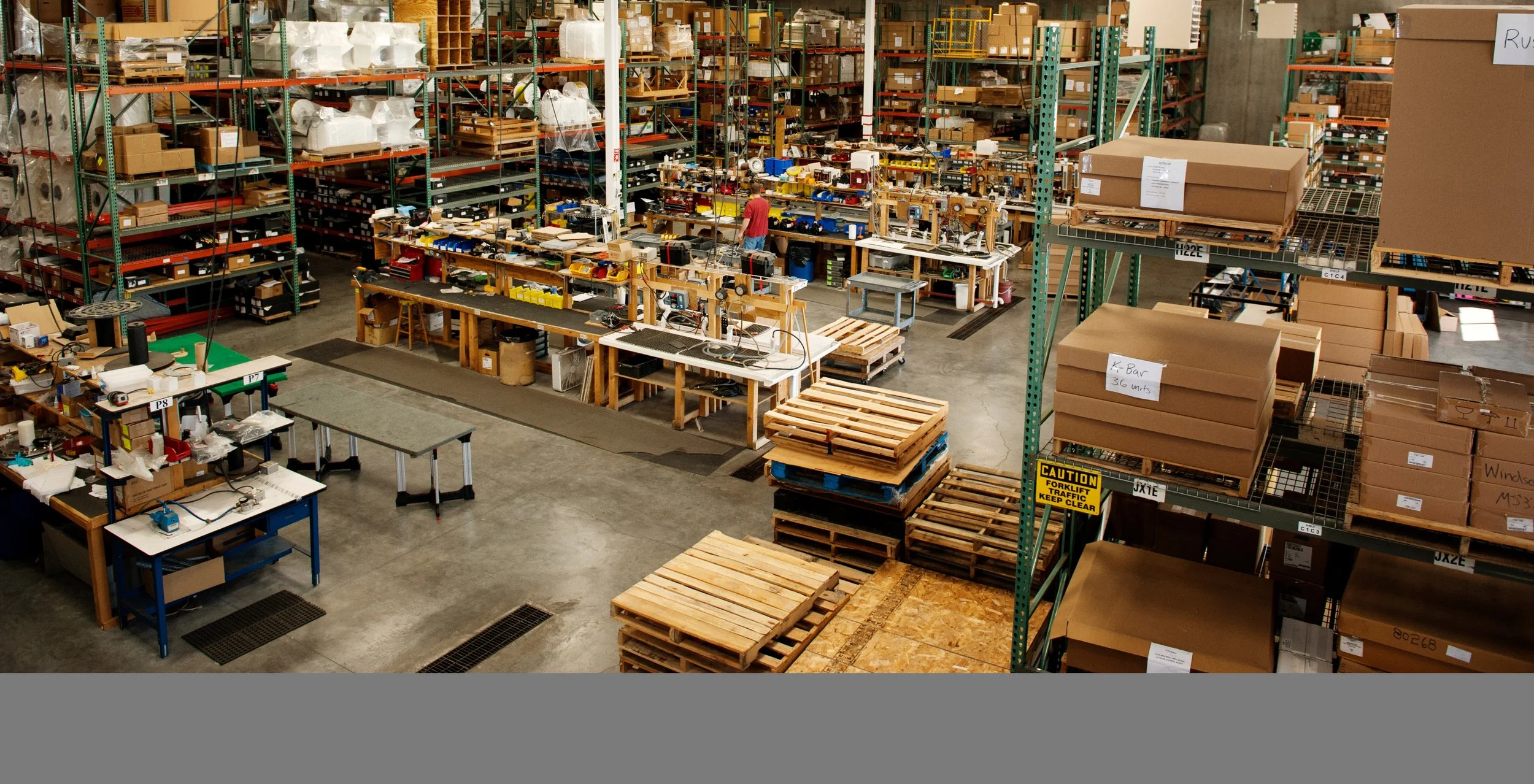 An ecommerce fulfillment warehouse with metal shelving filled with boxes and supplies, workstations with tools and electronics, and stacked wooden pallets.