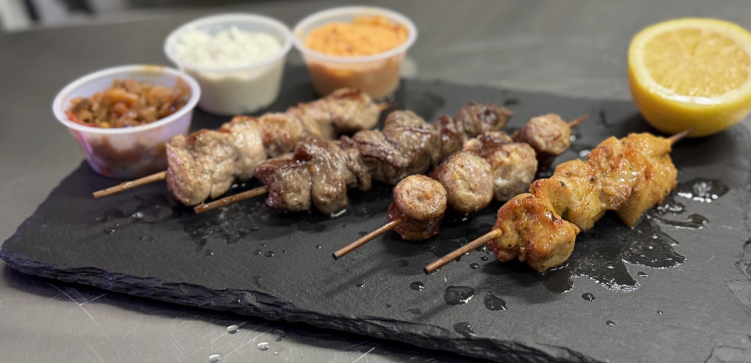 Three skewers of grilled meat on a black slate serving platter, with small bowls of sauces, lemon half, and sliced lemon in the background.