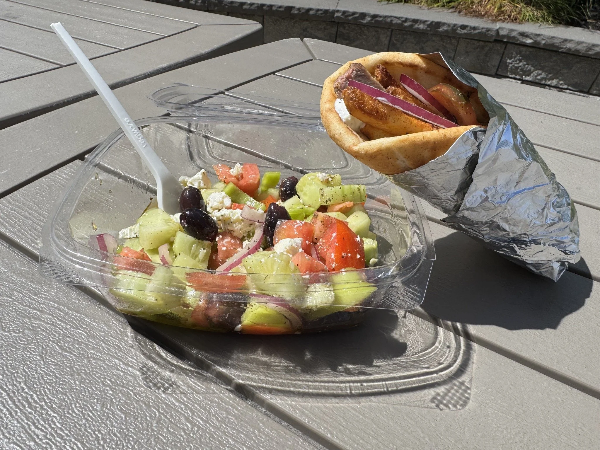 Fresh Greek salad with cucumbers, tomatoes, red onions, black olives, and feta cheese in a clear plastic container, paired with a gyro wrap filled with meat, onions, and tomatoes, on a gray outdoor table.