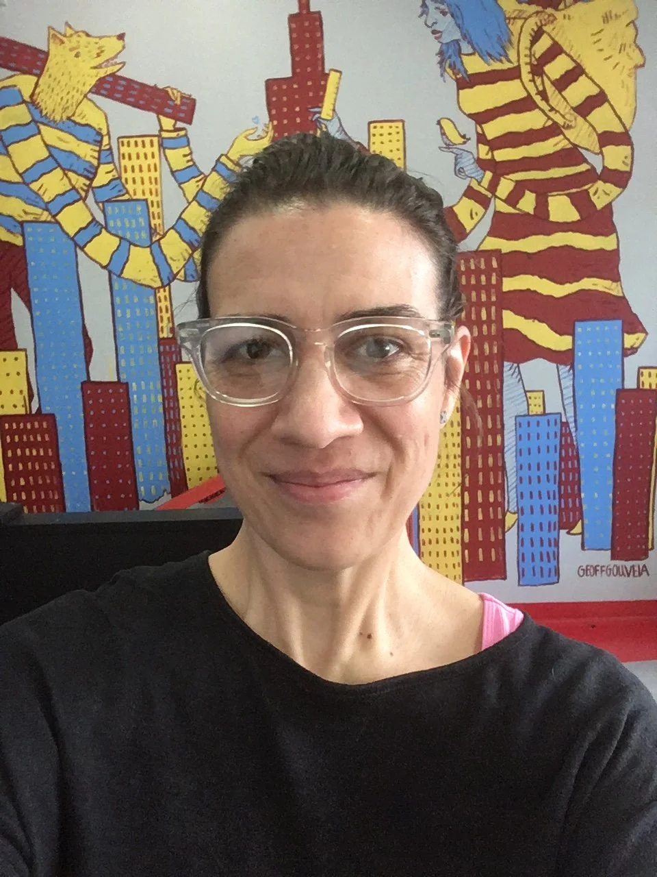a woman with fair skin and dark hair wearing glasses, in front of a colorful mural