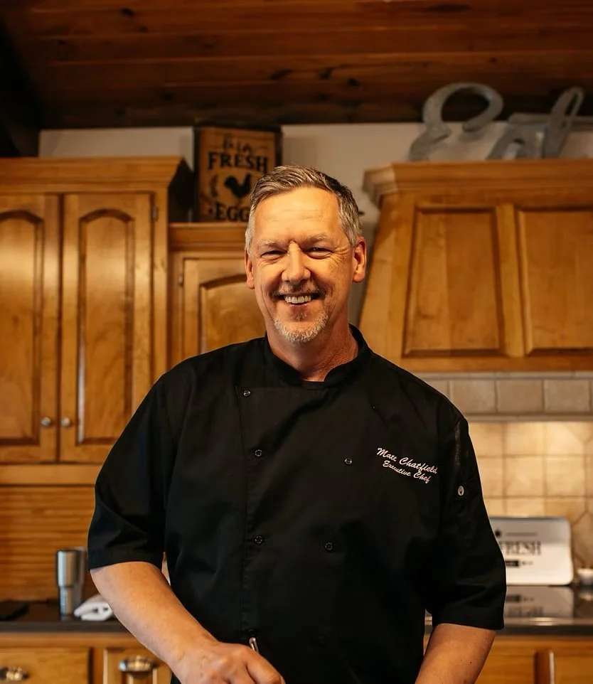 A man wearing a black chef coat smiling in a kitchen with wooden cabinets.
