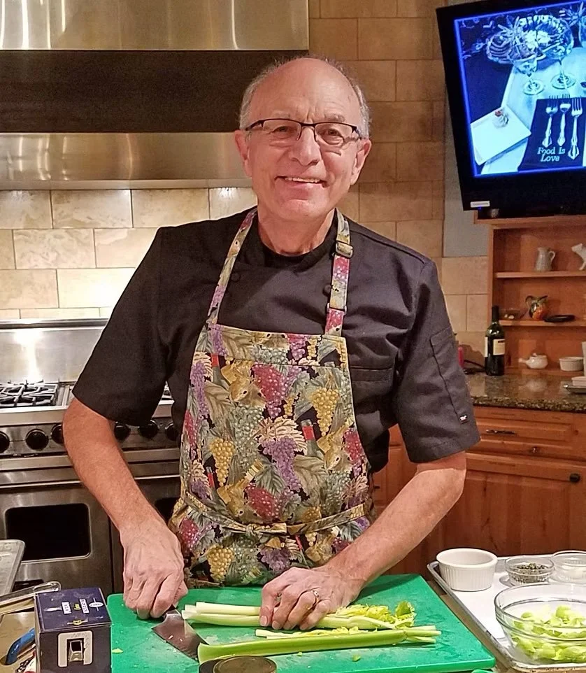 An older man with glasses, wearing a black chef's coat and a floral apron, standing in a kitchen chopping celery on a green cutting board.