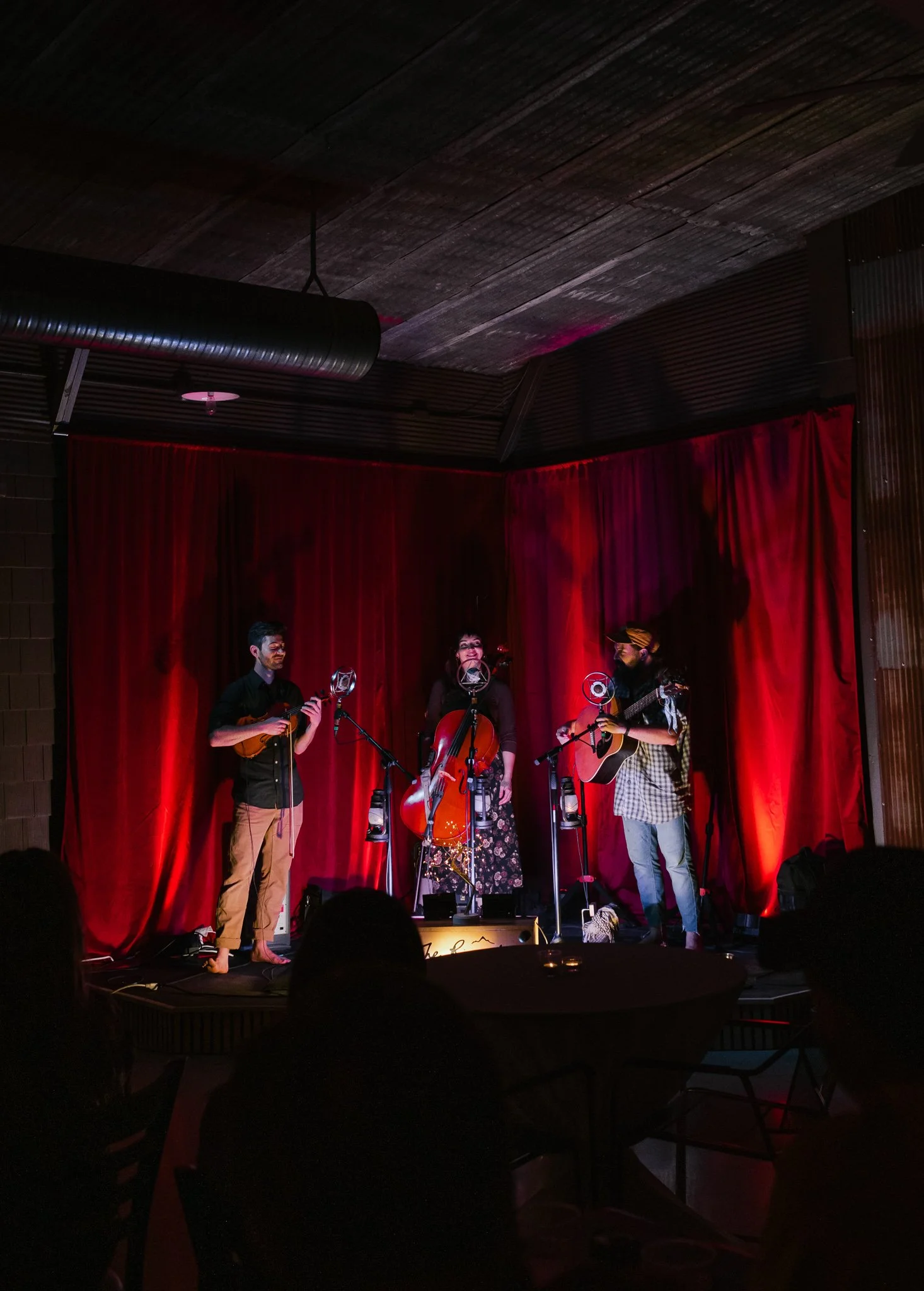 Three musicians performing on stage with red curtains in the background. They are playing string instruments and singing into microphones. The scene is in a dimly lit venue with an audience in front.
