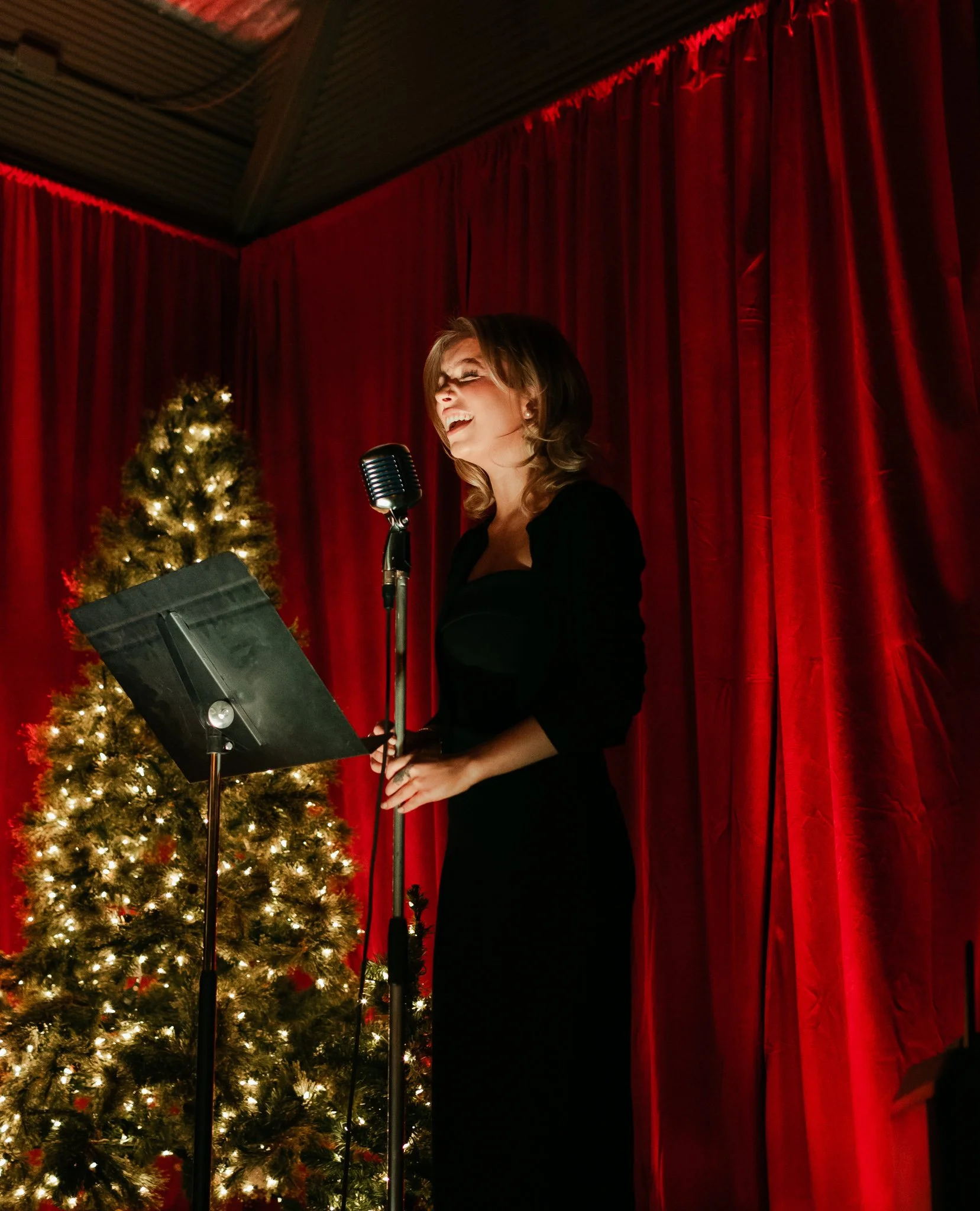 A woman singing or performing on stage with a microphone, standing in front of red curtains and a decorated Christmas tree with lights.