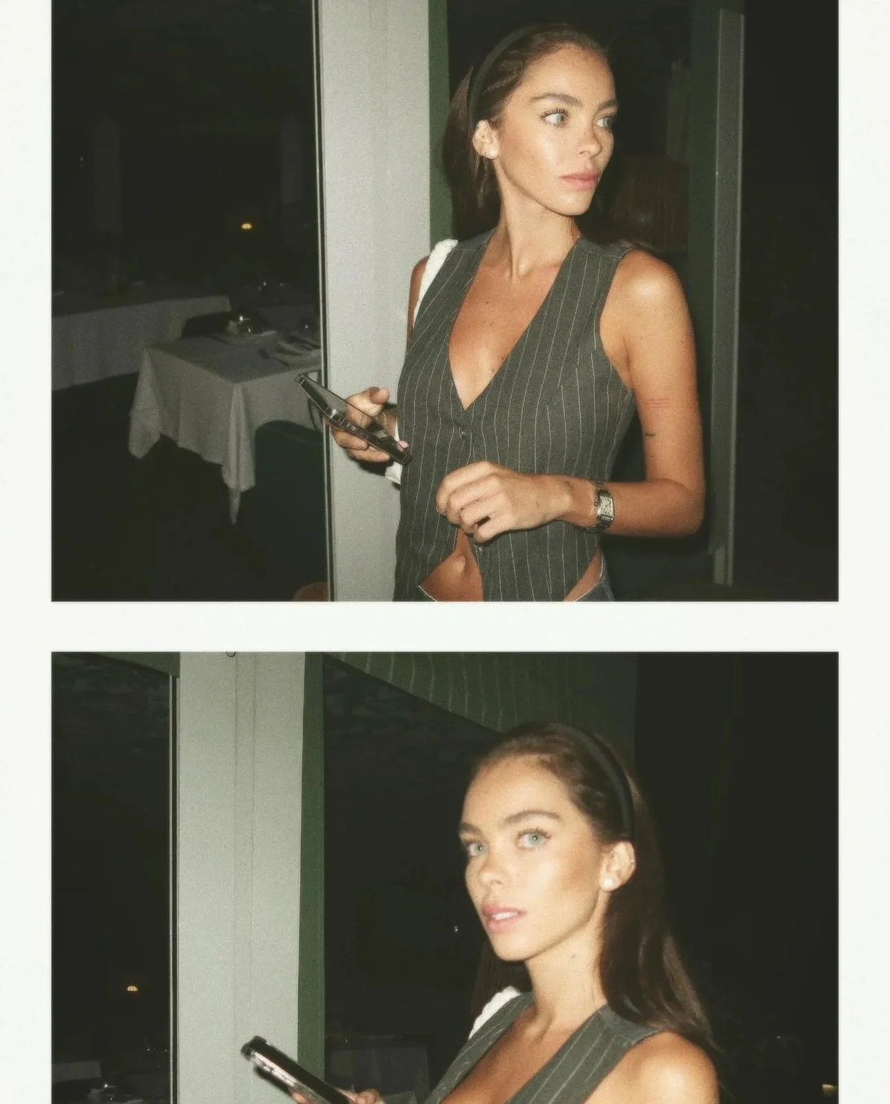 Mulher com cabelo castanho, vestindo roupa de cor cinza com listras brancas, segurando um telefone celular e usando máscara de proteção nos cabelos. Ela está em ambiente interno, possivelmente uma sala de jantar, com mesa coberta por toalha branca ao fundo.