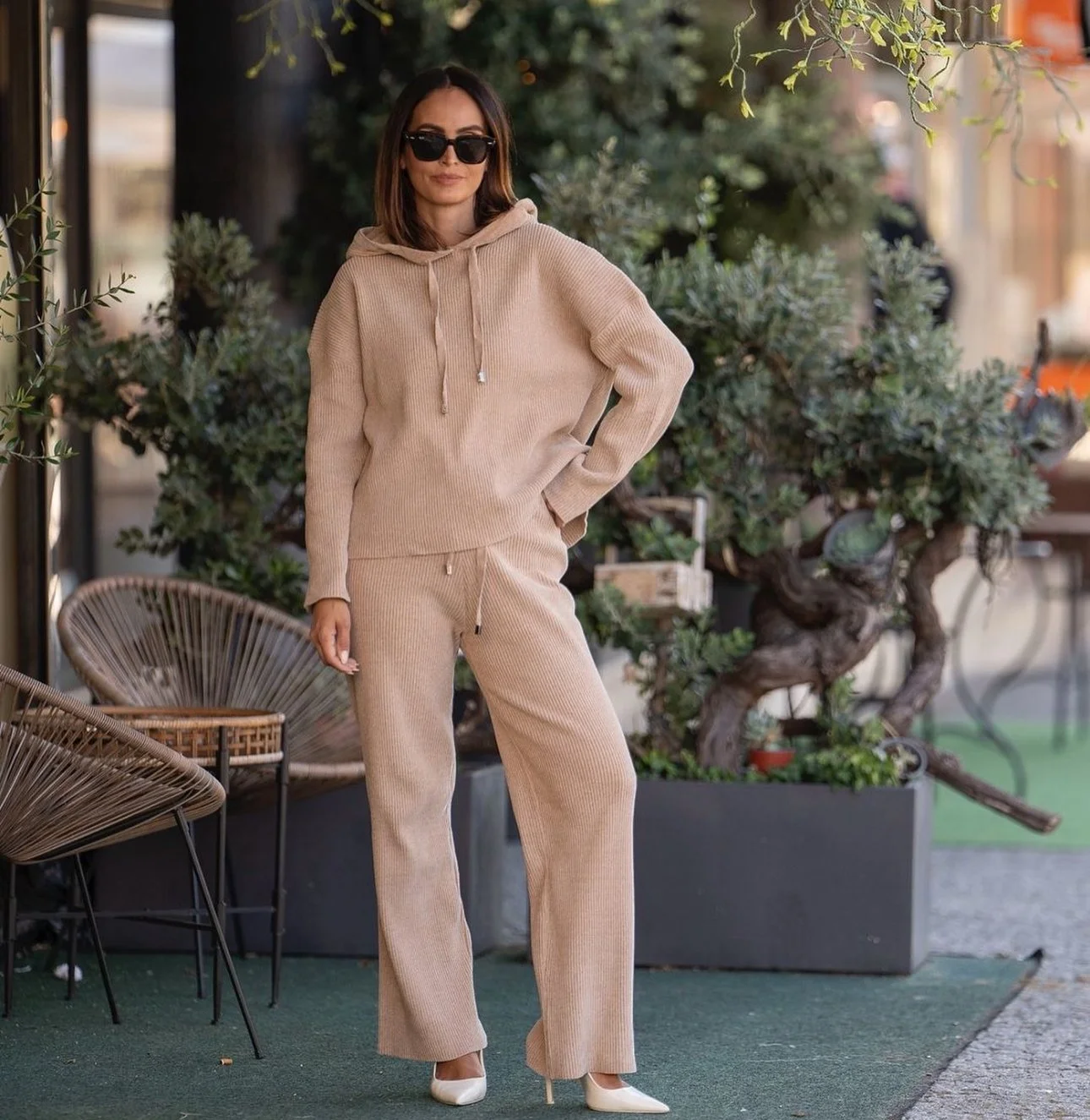 Mulher vestindo conjunto de moletom bege com capuz e calça de elástico, usando óculos escuros e sapatos brancos, posando ao lado de cadeiras de vime e plantas em um ambiente ao ar livre.