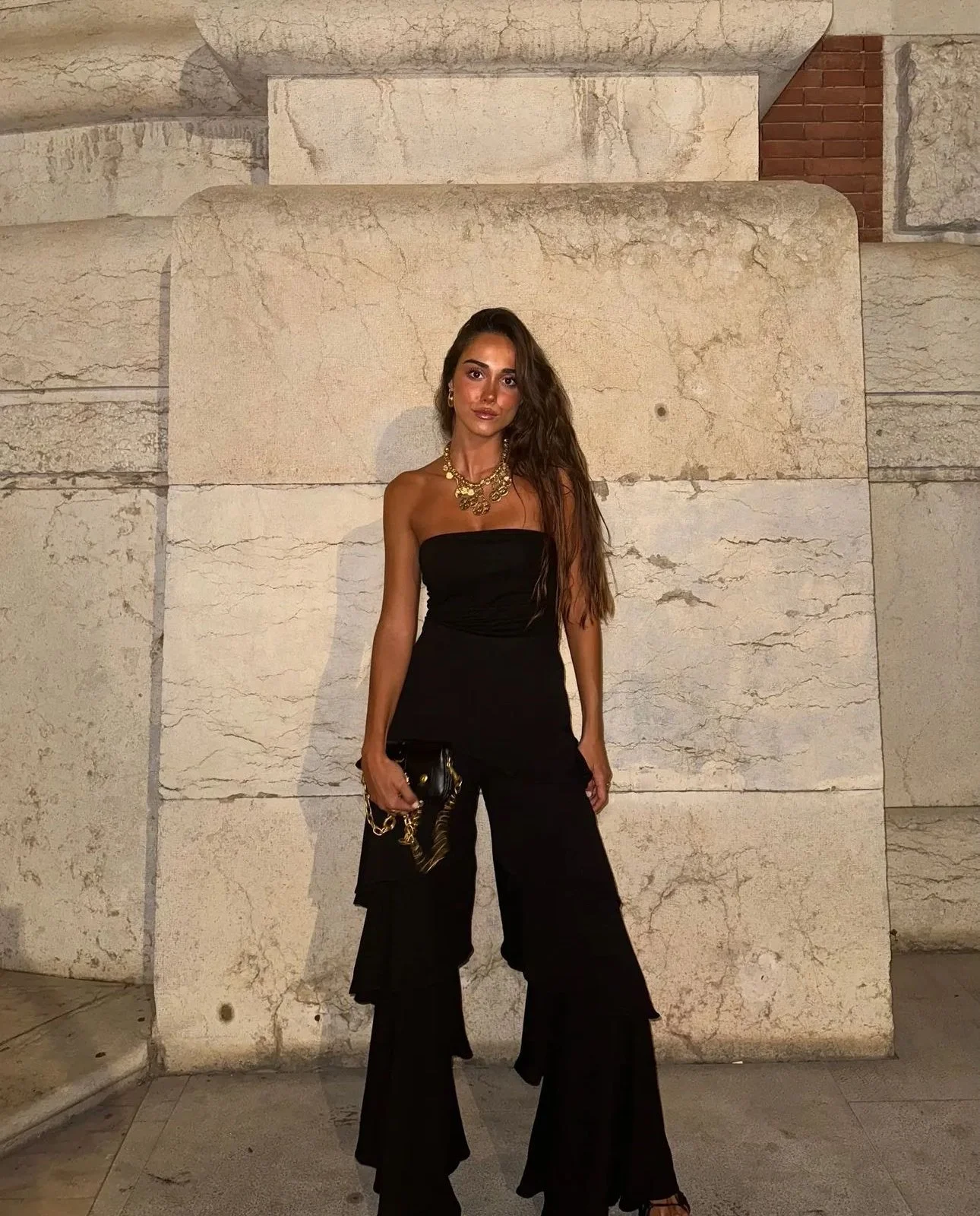 Mulher com cabelo castanho comprido, vestindo um macacão preto com pantalonas largas e uma joia dourada, segurando uma bolsa preta com corrente dourada.