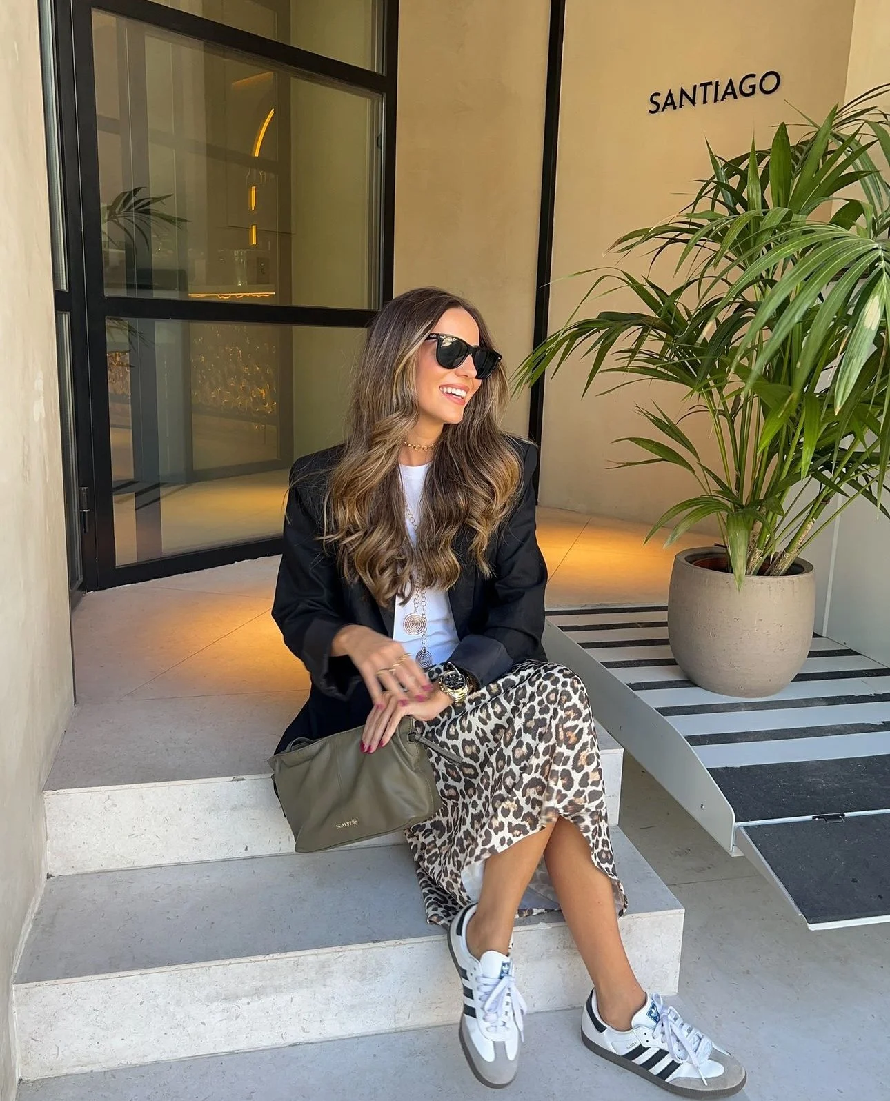 Mulher sorridente com cabelo cacheado usando óculos escuros, brincando com um relógio, sentada em degraus de uma entrada com uma planta ao lado e a palavra 'SANTIAGO' na parede de fundo