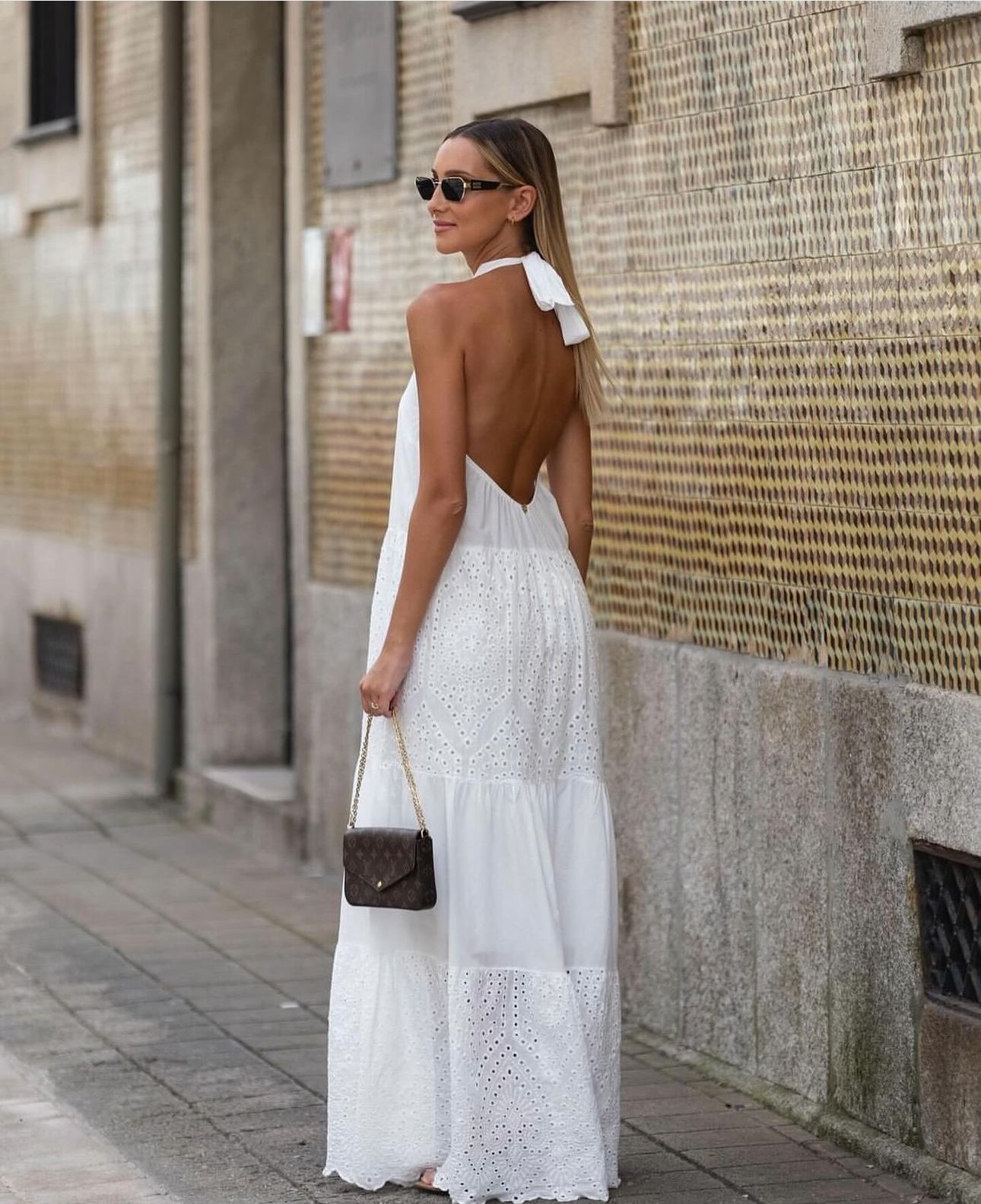 Mulher com vestido branco longo com costas abertas, segurando uma bolsa marrom com alça de corrente, caminhando na rua.