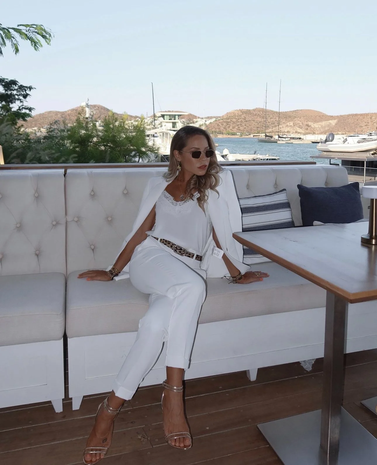 Mulher elegante sentada em sofá branco ao ar livre com vista para o mar e barcos, usando óculos escuros, roupa branca, acessórios e sapatos de salto.