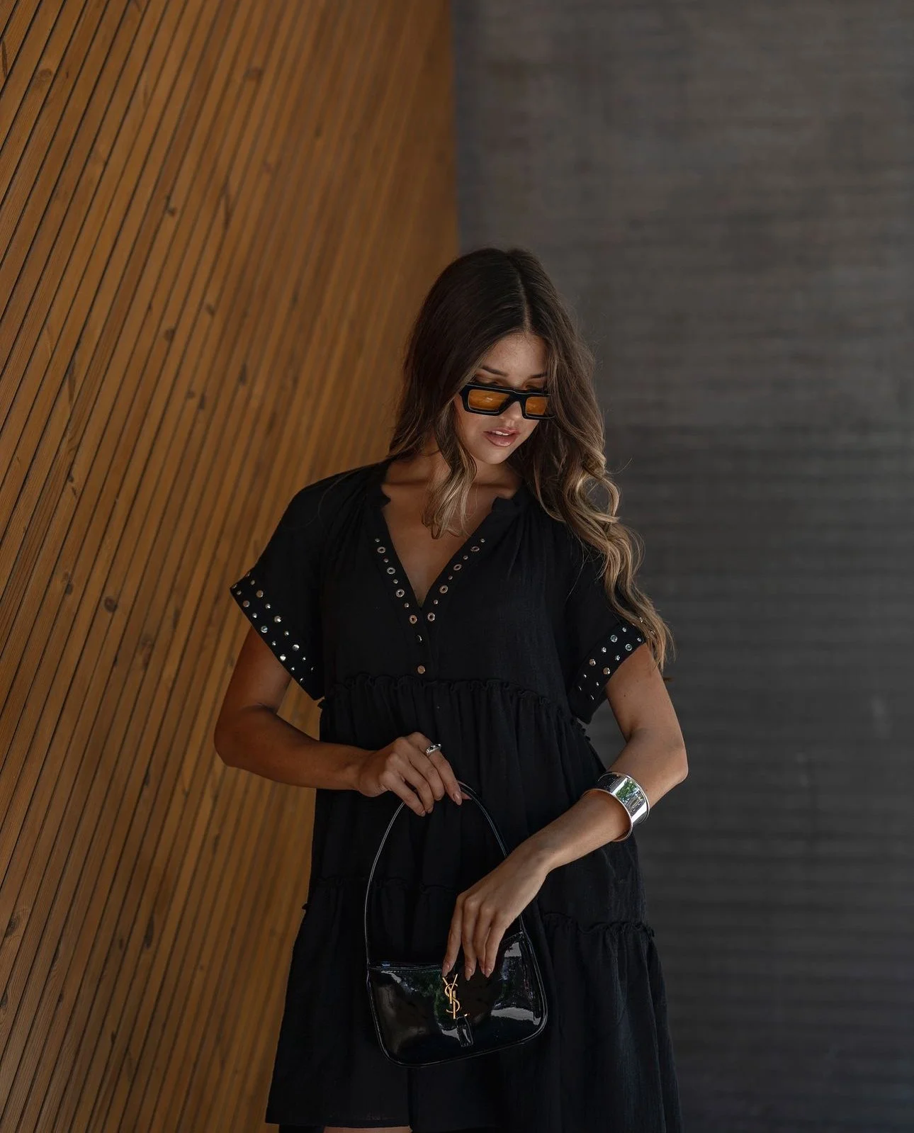 Mulher com cabelo castanho, cabelo longo, usando óculos escuros e vestido preto com detalhes de metal, segurando uma bolsa preta com logotipo dourado, em ambiente interior com paredes de madeira e parede cinza.