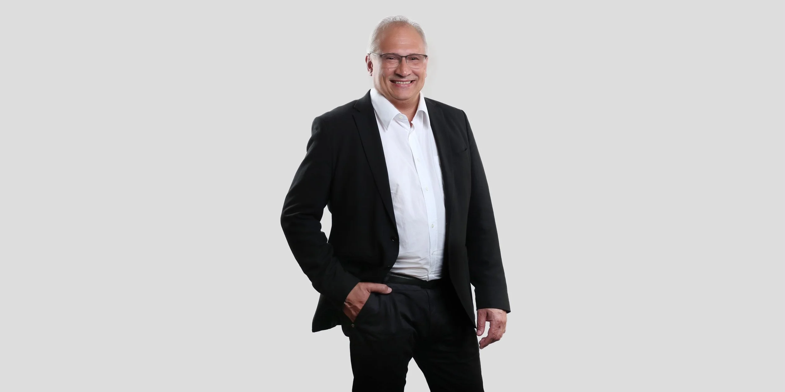 Ein älterer Mann in einem schwarzen Anzug mit weißem Hemd, lächelnd, auf grauem Hintergrund