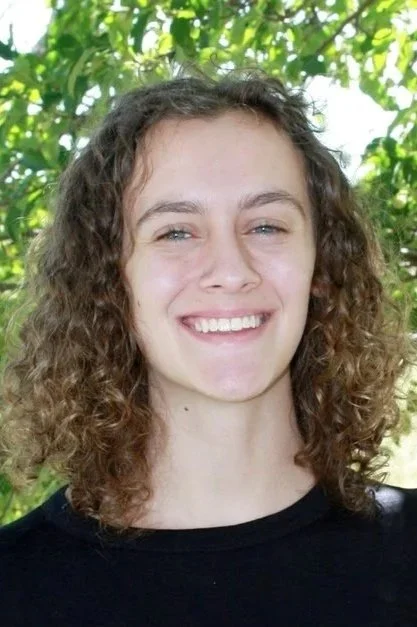 A young woman with curly brown hair smiling outdoors in front of green foliage, wearing a black shirt.