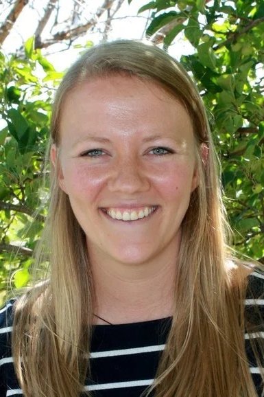 A young woman with long blonde hair smiling outdoors, with green foliage in the background.