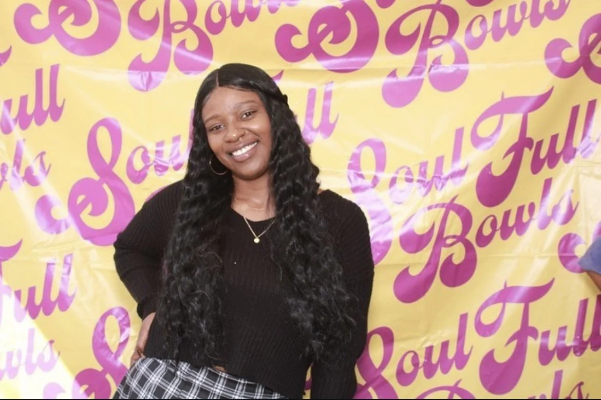 Smiling woman with long, wavy black hair wearing a black sweater and a plaid skirt standing in front of a yellow background with pink cursive text that says 'You Full' repeatedly.