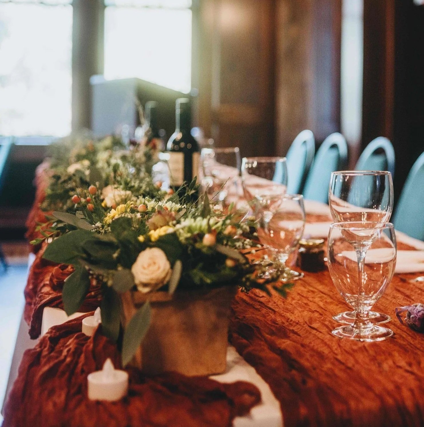 A beautifully decorated dining table with a floral centerpiece, wine glasses, and table settings in a cozy, warmly lit room with natural light coming through windows.
