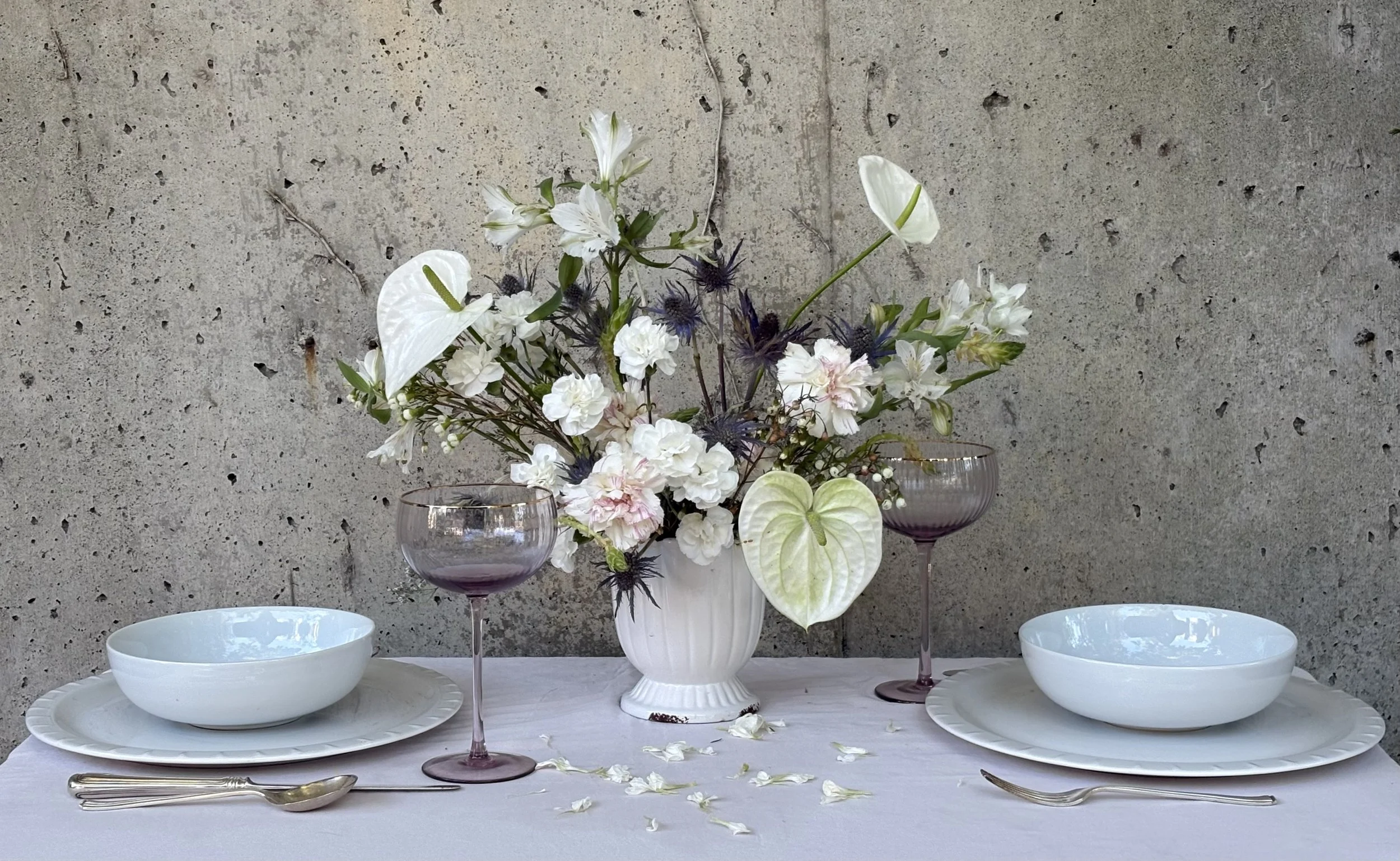 Elegant table setting with white bowls, silverware, and two purple wine glasses, featuring a floral centerpiece with white, blush, and cream flowers, anthuriums, and greenery against a concrete wall background.