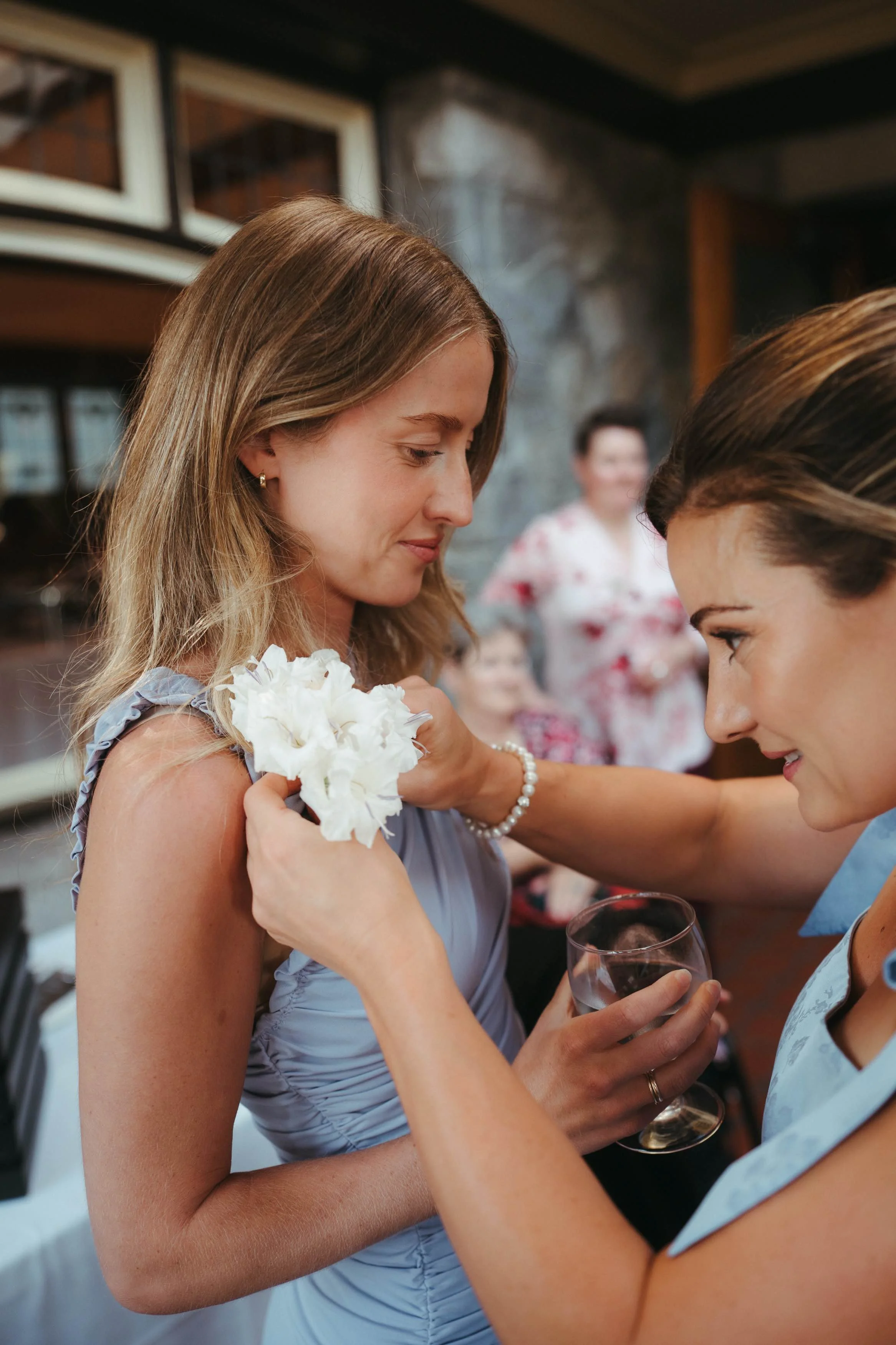 A woman in a light purple dress receiving a corsage from another woman in a blue dress at a gathering, with several other women in the background.