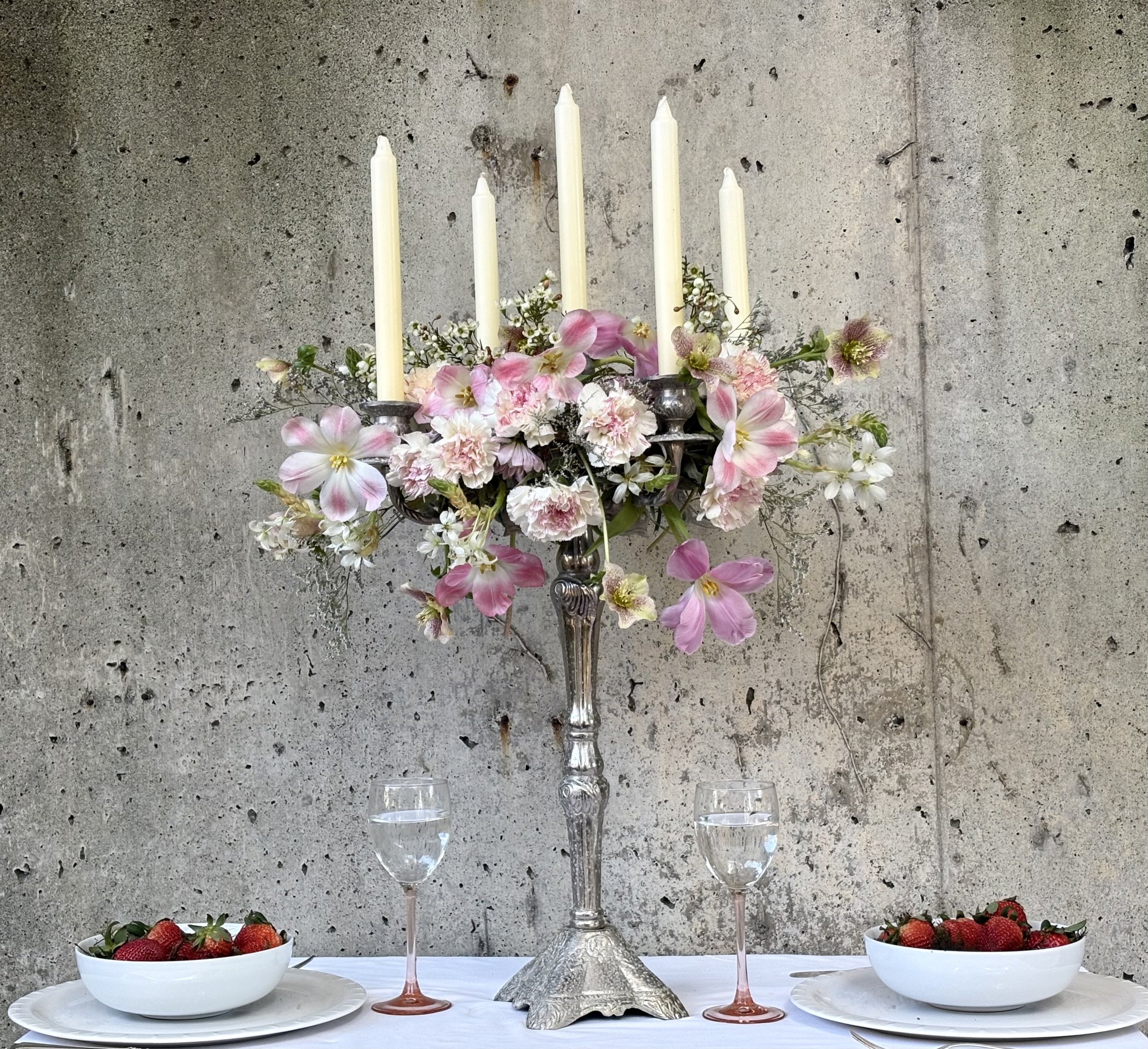 A table setting featuring a large floral candelabrum with pink and white flowers, holding five white candles, flanked by two bowls of strawberries, two glasses of water, with a plain concrete wall in the background.