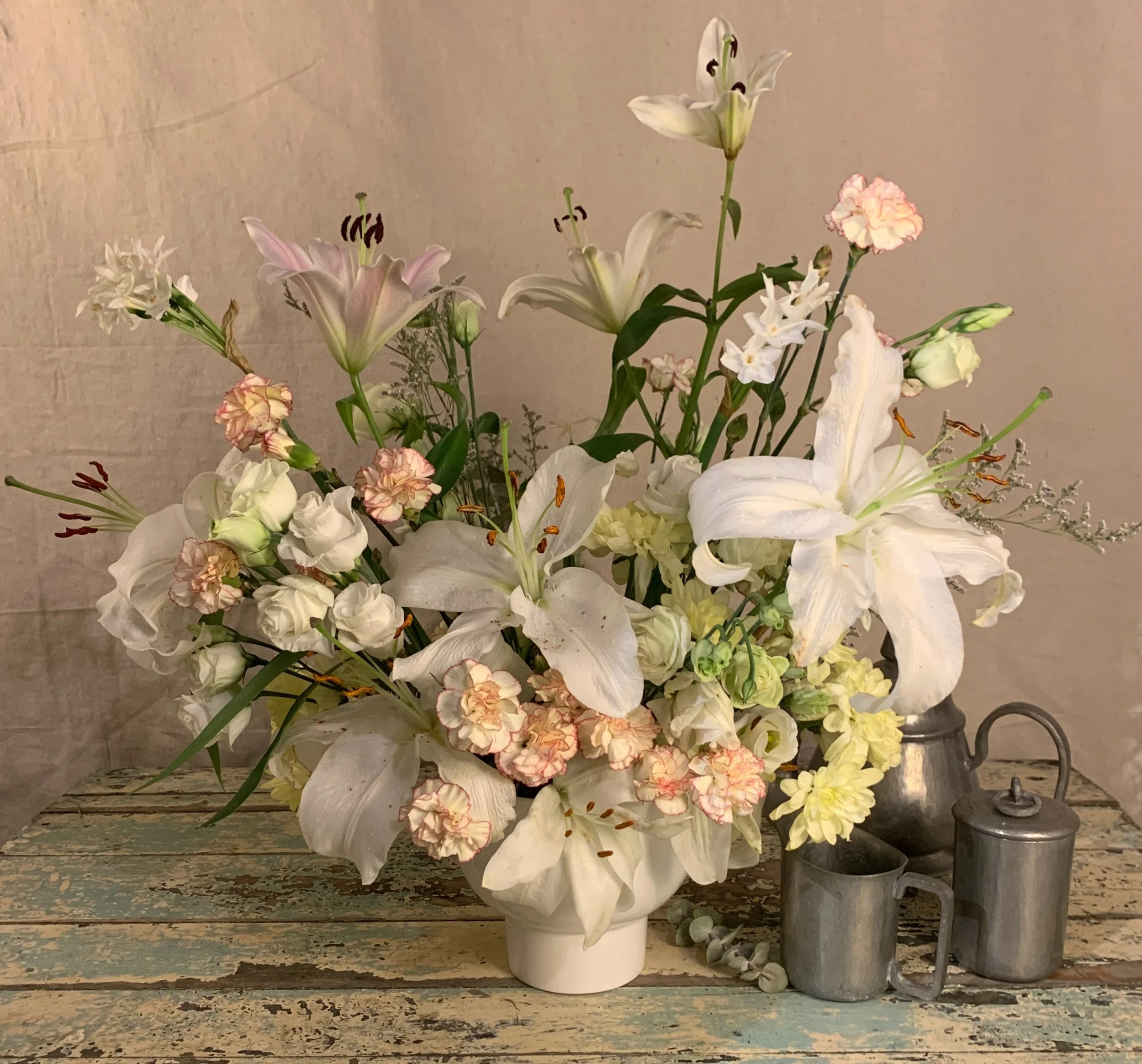 A large bouquet of white lilies, pink and white carnations, and smaller white and pink flowers in a white vase, on a rustic wooden table with metal teapots and cups.