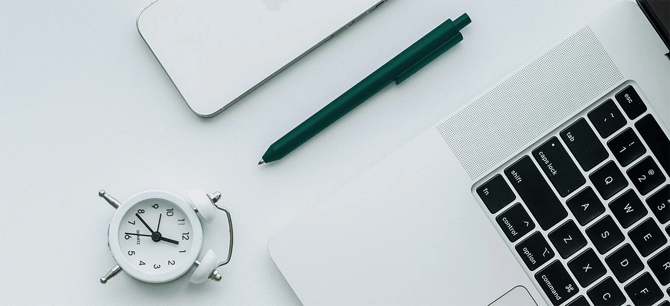A green pen, a MacBook, an iPhone, and a clock lay on a white desktop