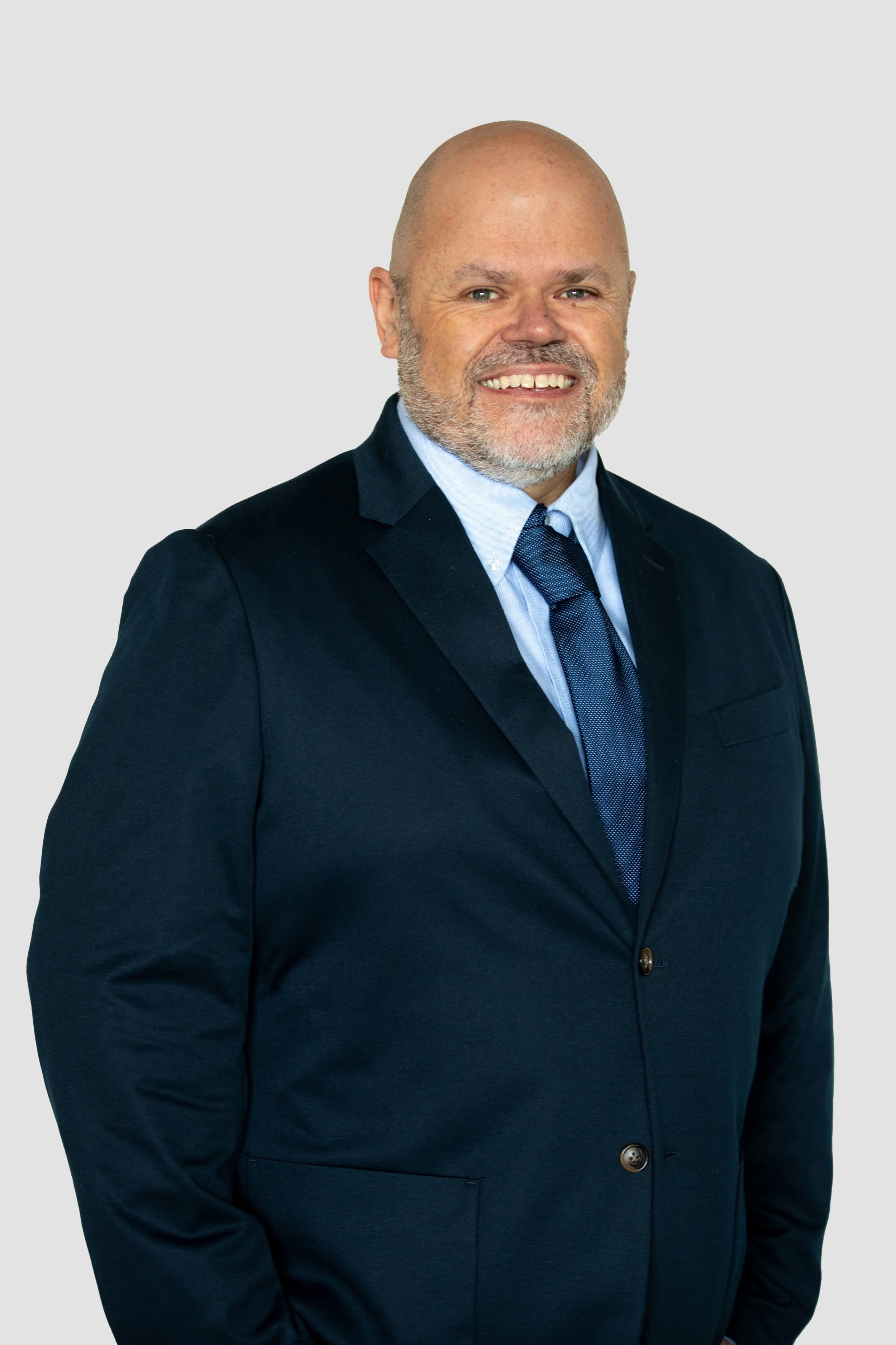 Ken Higginson dressed in a dark suit with a blue shirt and tie, smiling against a plain light background.