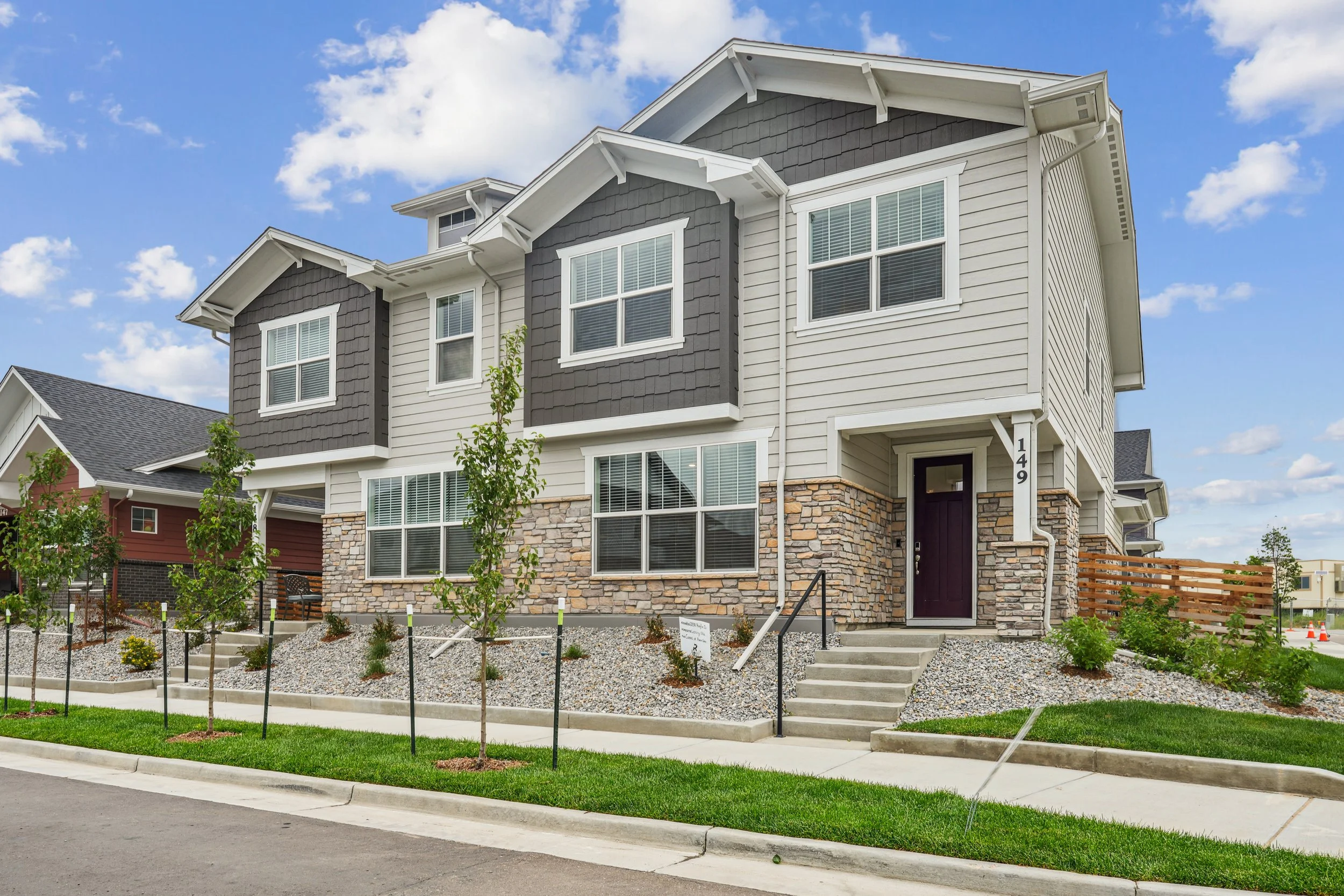 A modern multi-story home for rent in Brighton, CO with white siding, gray accents, and stone foundation, featuring multiple windows and a purple front door, situated on a landscaped lot with young trees and a sidewalk.