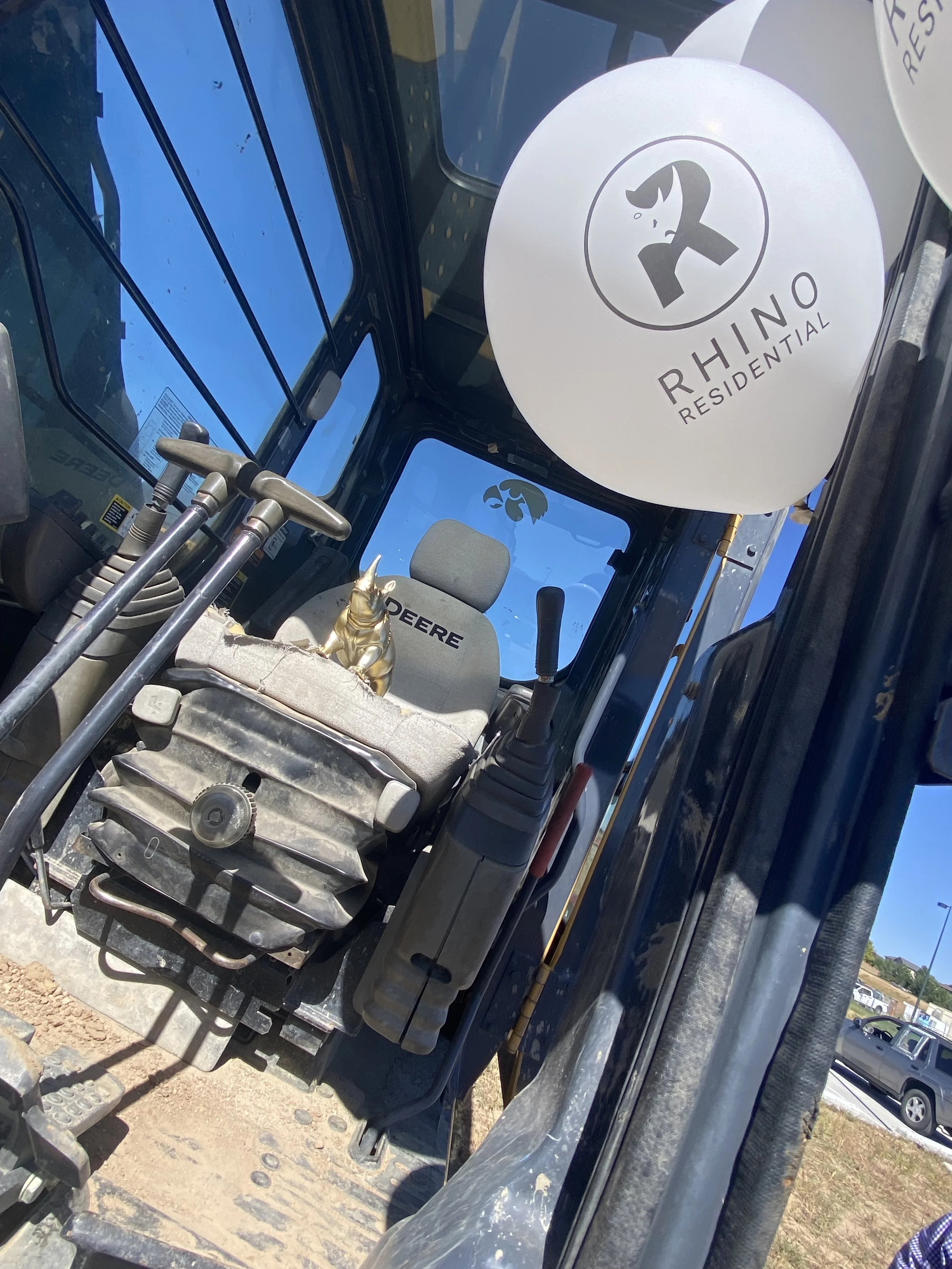 View of the front interior of a John Deere construction or agricultural vehicle, with a beige seat labeled 'John Deere,' controls, and two Baietti balloons with 'Rhino Residential' branding outside the windows.