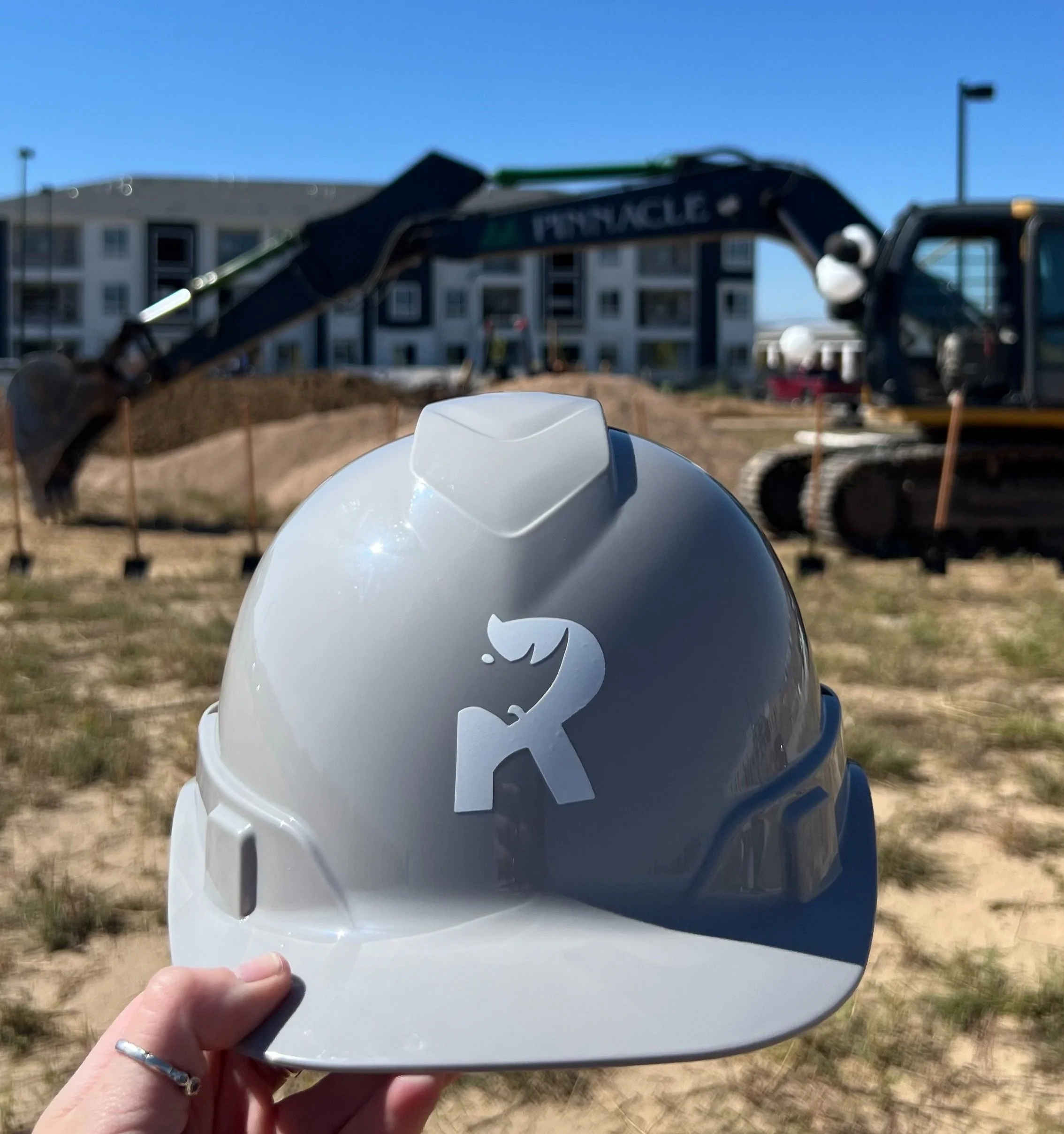 Person holding a white construction helmet with rhino residential logo, at a construction site with an excavator and building in the background.