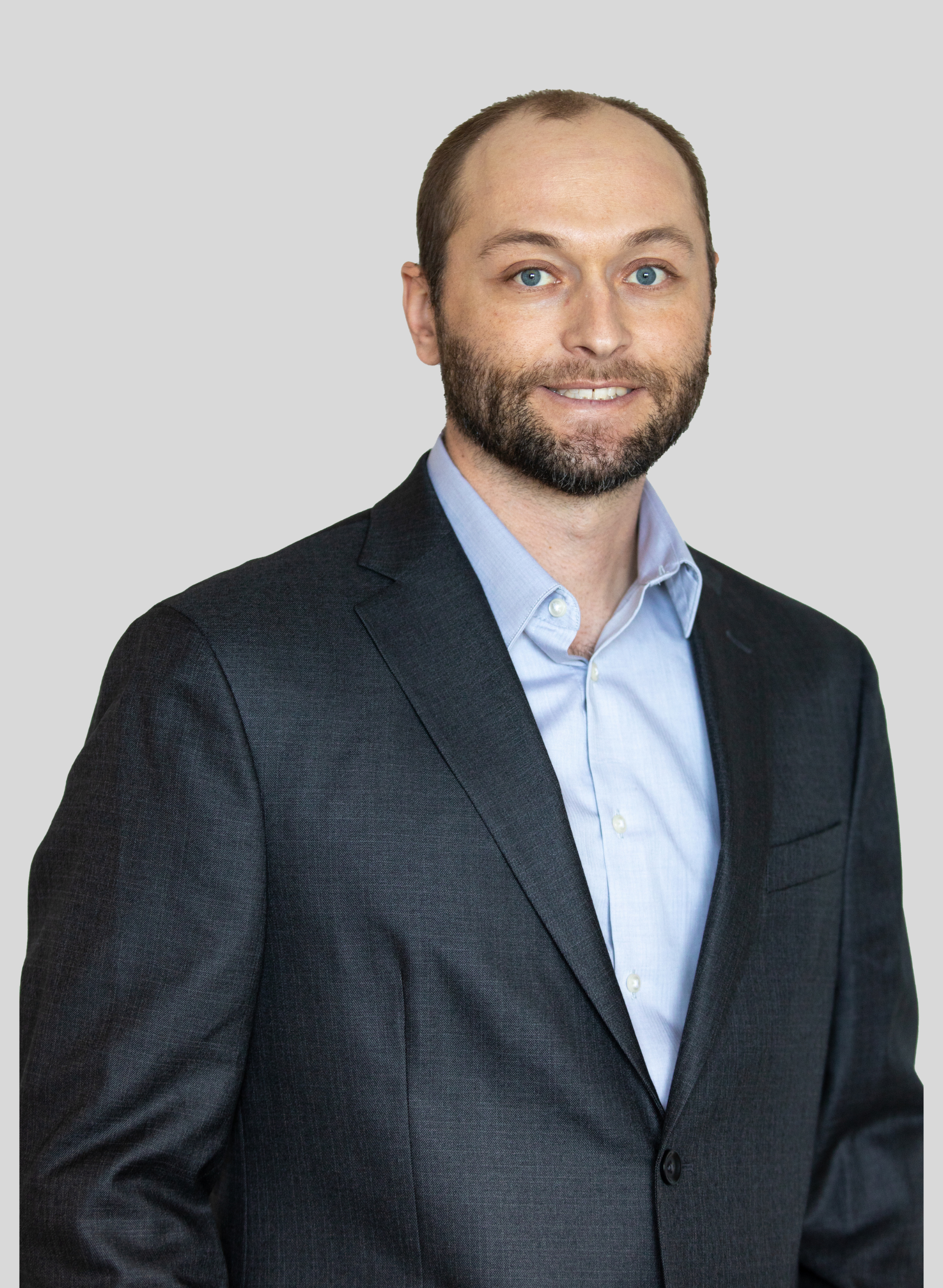 Zach Stamp Professional headshot wearing a navy blazer and a light blue dress shirt, standing against a plain light gray background.