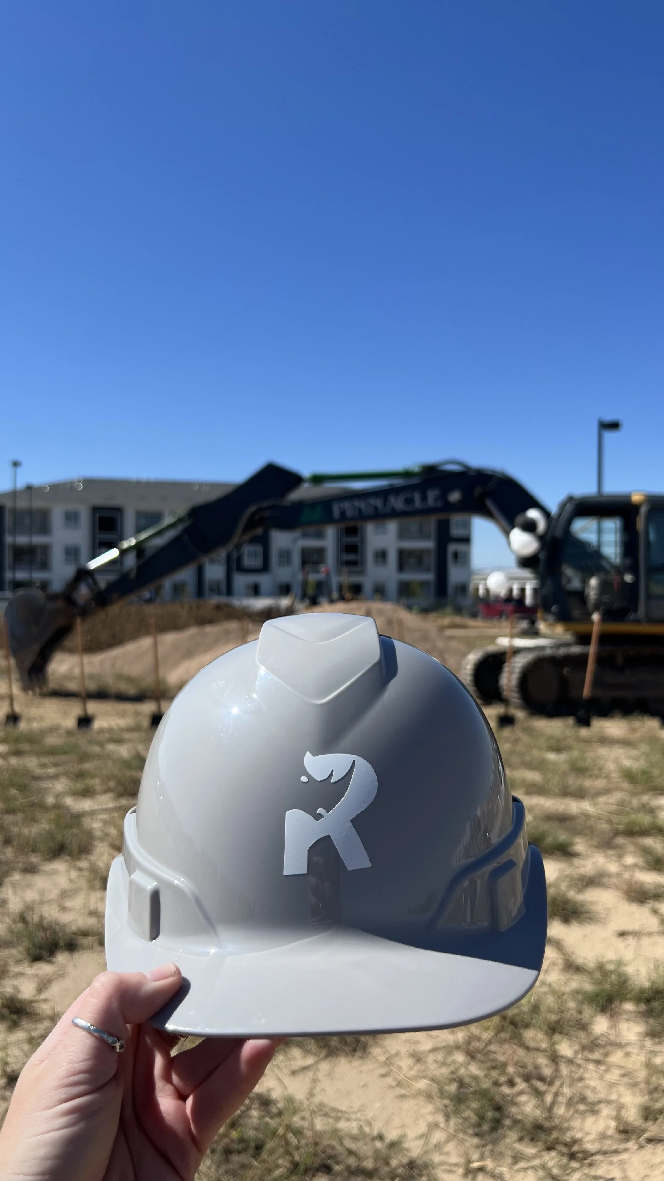 Person holding a white construction helmet with a black number 2 logo in front of a construction site with a bulldozer and an excavator.