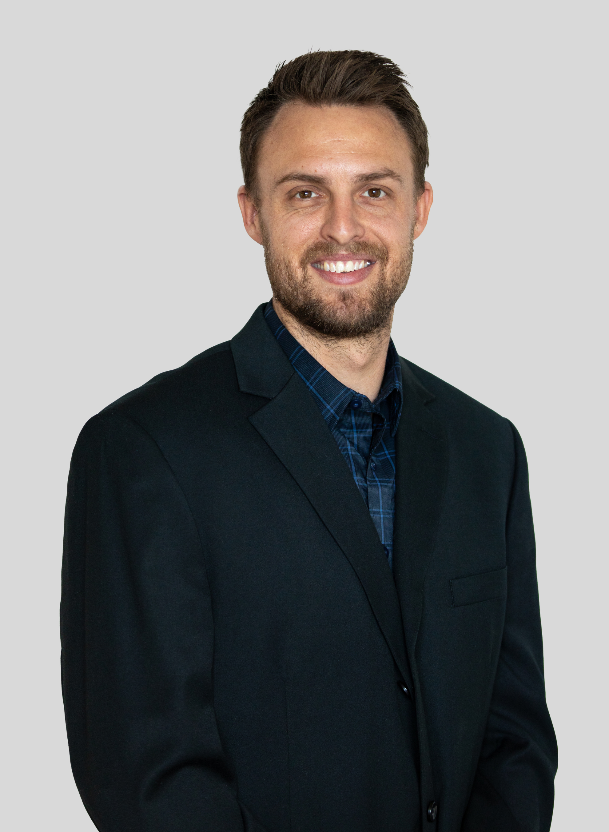 Scott Arpin with brown hair and a beard wearing a black blazer and blue checkered shirt, standing against a plain gray background.