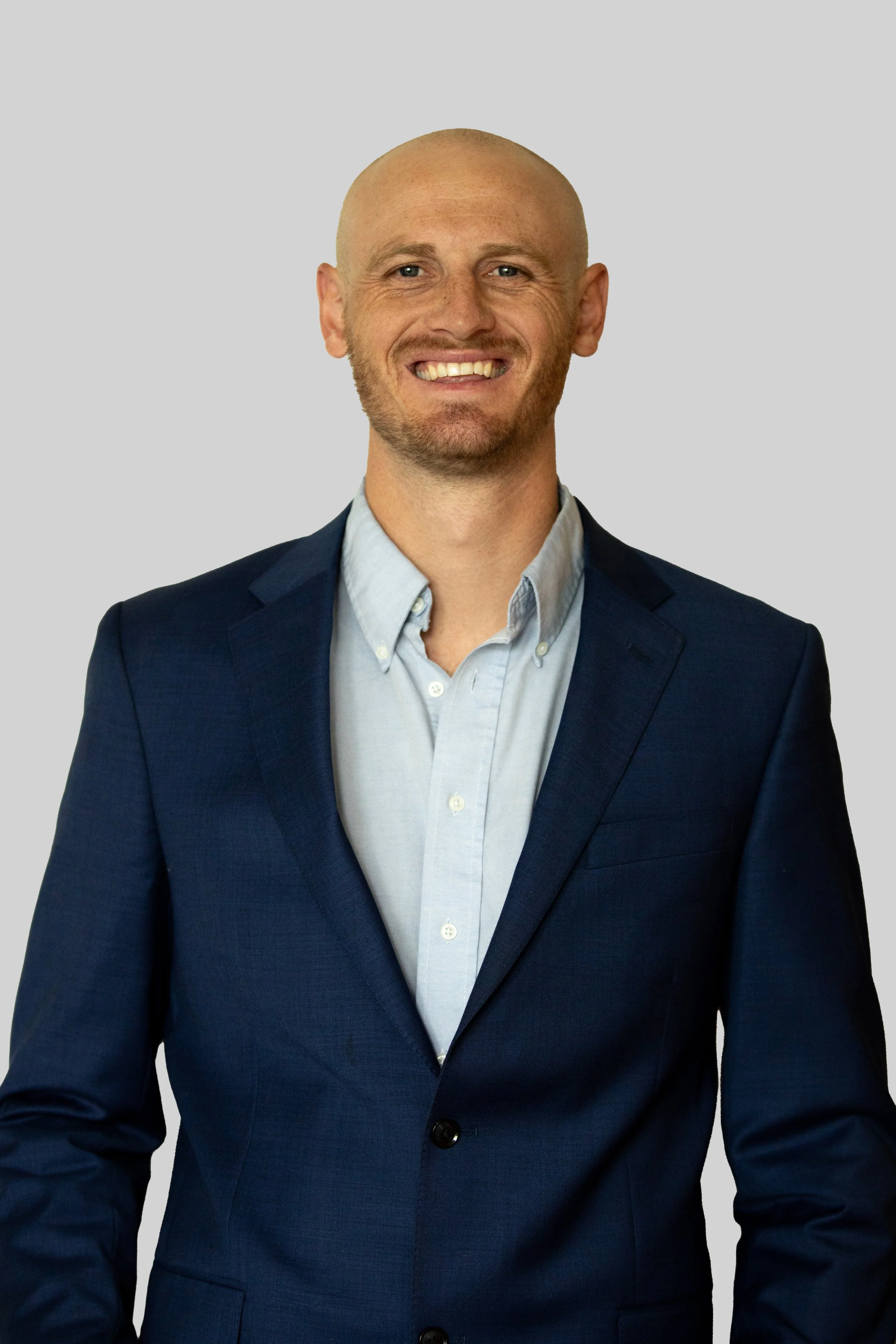 Xander Lambert wearing a blue blazer and a light blue button-up shirt, smiling at the camera against a plain gray background.