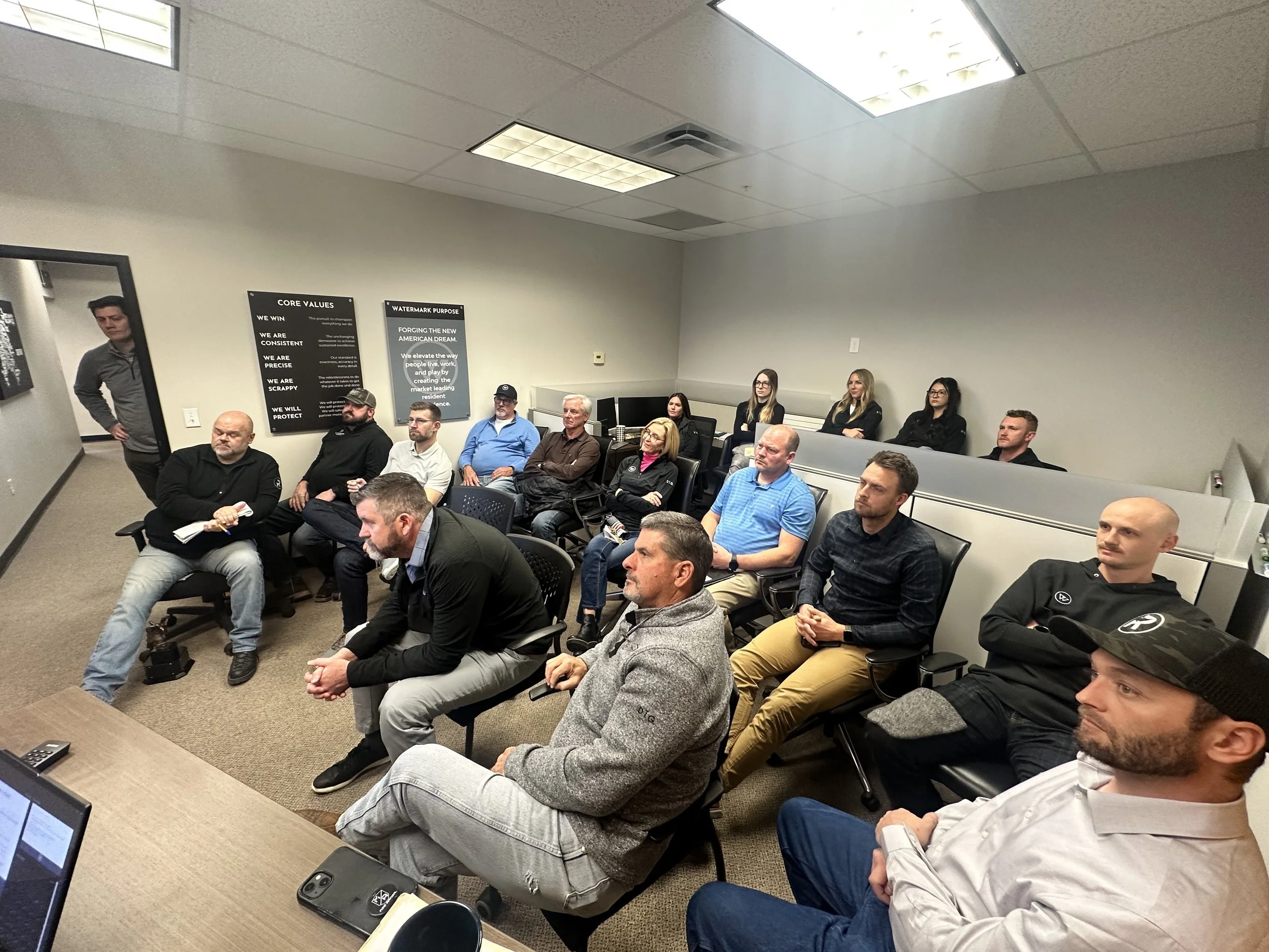 A group of people attending a meeting or seminar in a conference room. The room has beige carpeting, gray walls, and ceiling lights. Attendees are seated in chairs arranged in rows, some taking notes and listening attentively.