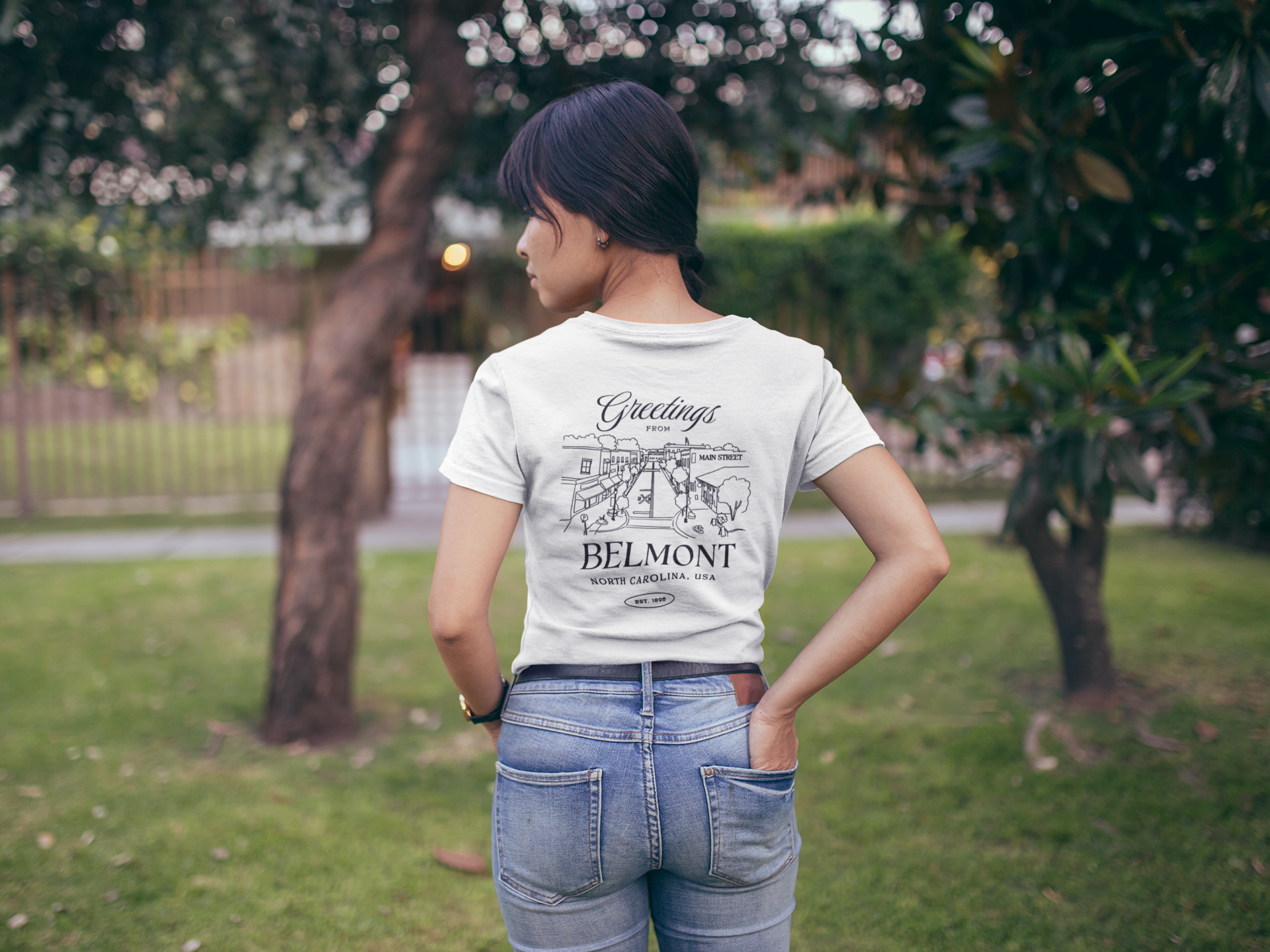 Person wearing a white t-shirt with "Greetings from Belmont North Carolina, USA" text and illustration, standing outdoors in a park.