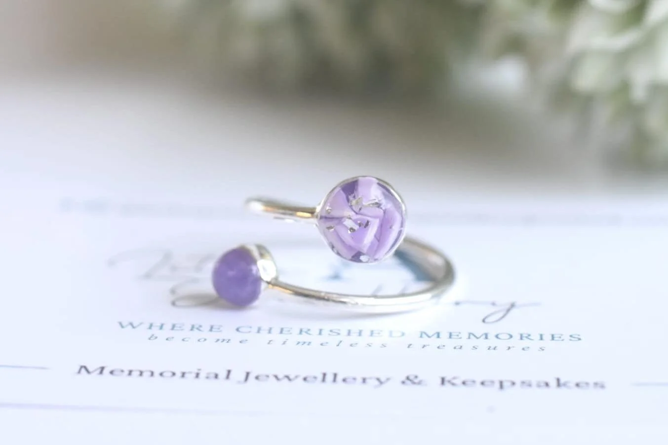 Two silver rings with purple glass or gemstone orbs resting on a paper that reads 'WHERE CHERISHED MEMORIES become treasures' and 'Memorial Jewellery & Keepsakes' with blurred background.