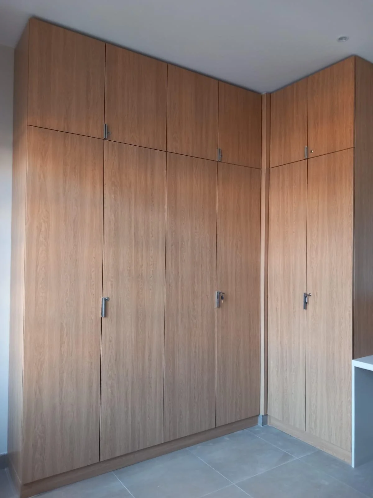 Grand placard en bois avec plusieurs portes, situé dans un coin d'une pièce, au sol en carreaux gris.