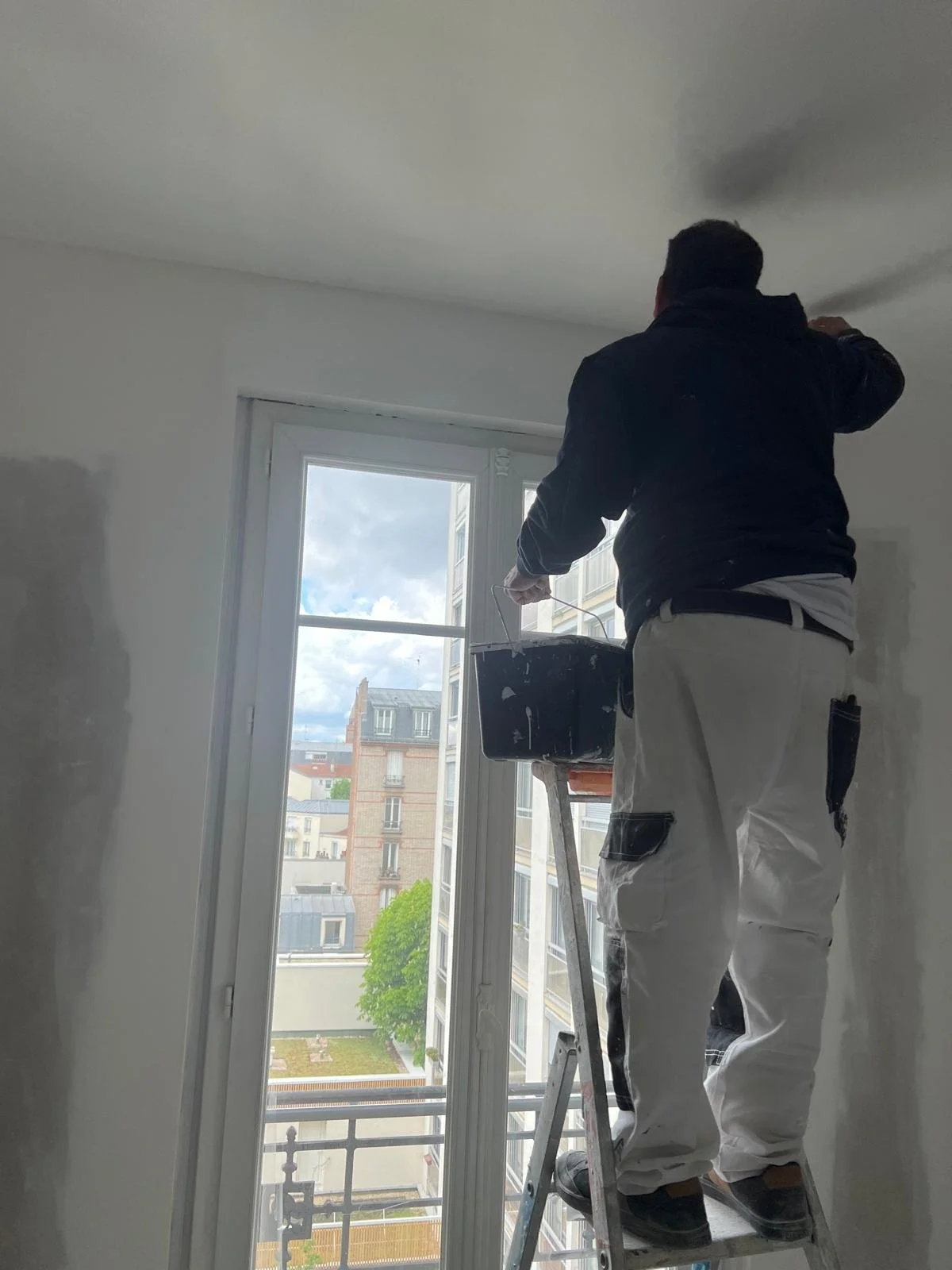 Un homme en train de peindre le plafond d'une pièce depuis une échelle, avec une vue sur un bâtiment et un ciel nuageux à travers une grande fenêtre.