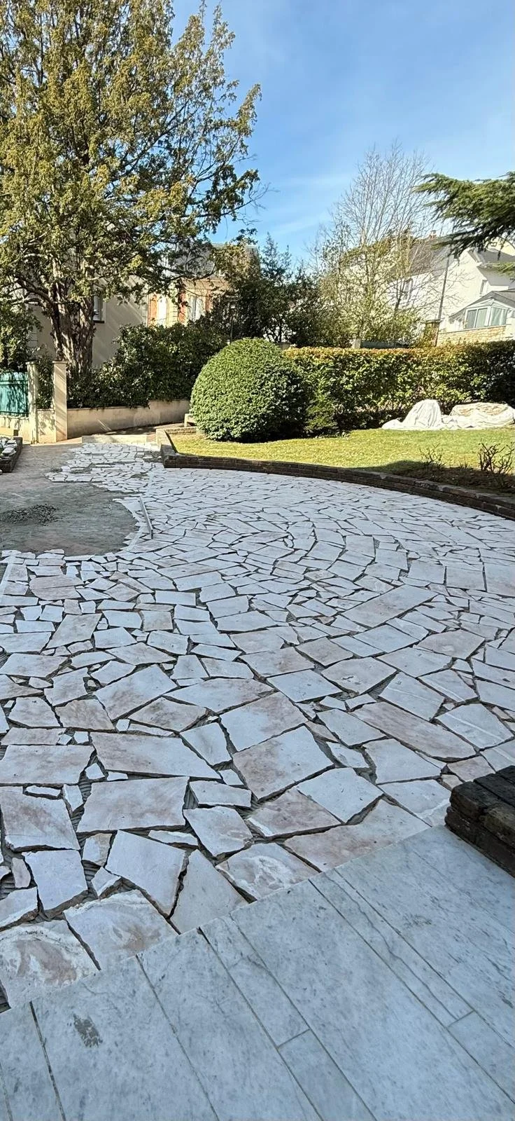 Un chemin pavé en pierre dans un jardin avec des arbustes et des arbres sous un ciel bleu.