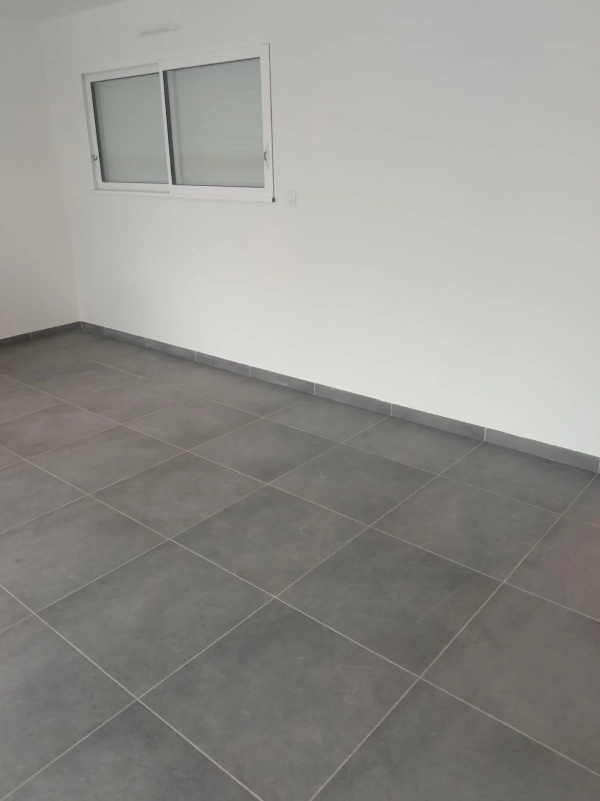 Pièce vide avec mur blanc, fenêtre à double vitrage et sol en carreaux gris.
