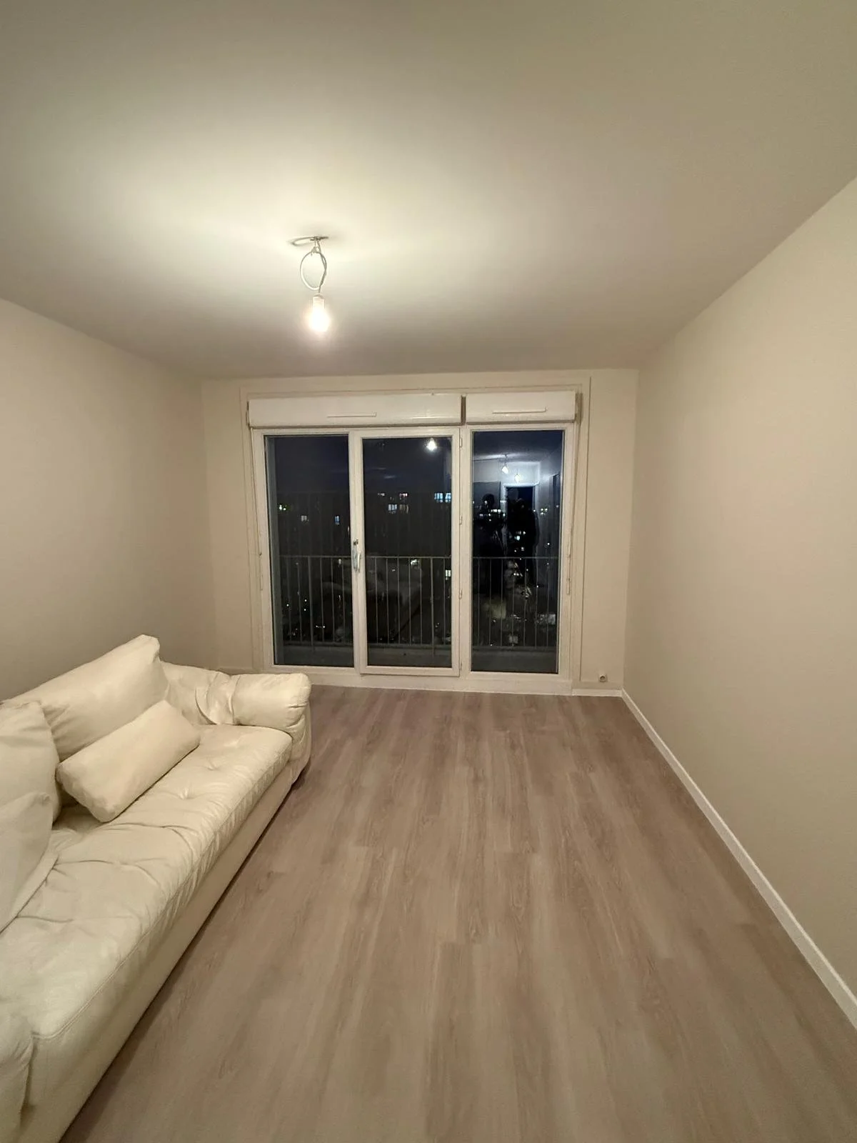 Salon vide avec un canapé blanc et une grande fenêtre donnant sur une ville la nuit.