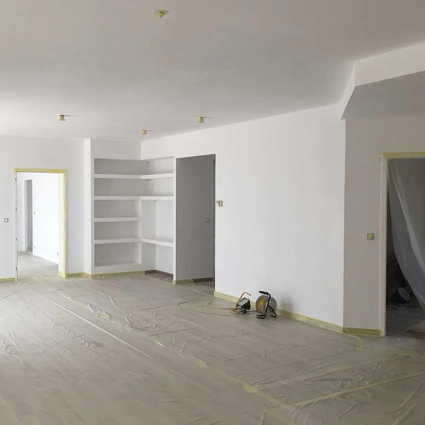 Intérieur d'une pièce en cours de rénovation avec murs et plafond blanc, rayures de protection sur le sol, étagères encastrées sur un mur, et équipements électriques posés par terre.