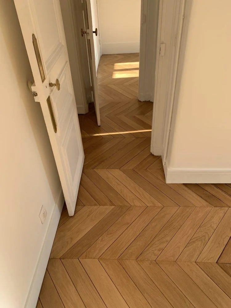 Un couloir intérieur avec un sol en parquet en chevrons, une porte gauche entrouverte et une lumière naturelle entrant par une fenêtre.