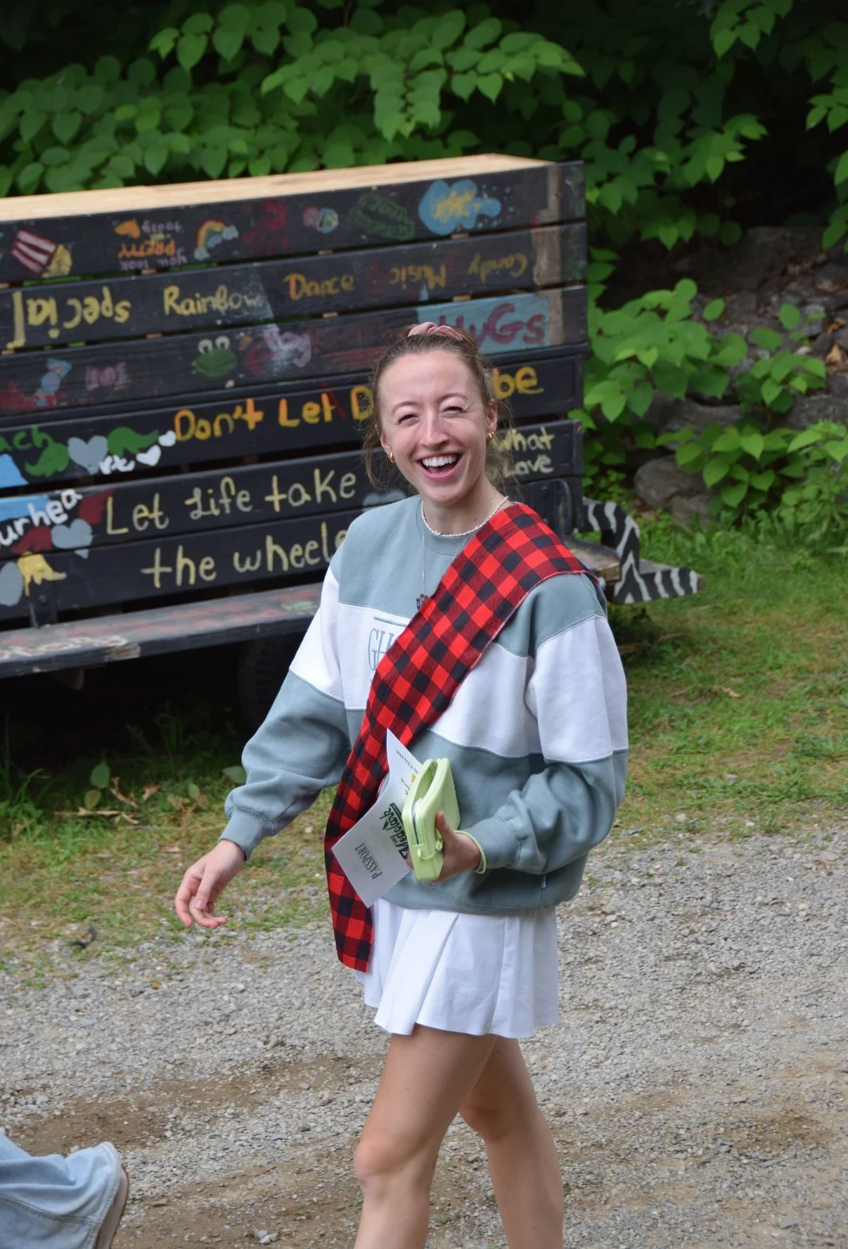 A young woman with a big smile, wearing a casual sweatshirt and a white skirt, holding papers and a green wallet, standing outdoors near a colorful painted bench and lush greenery.