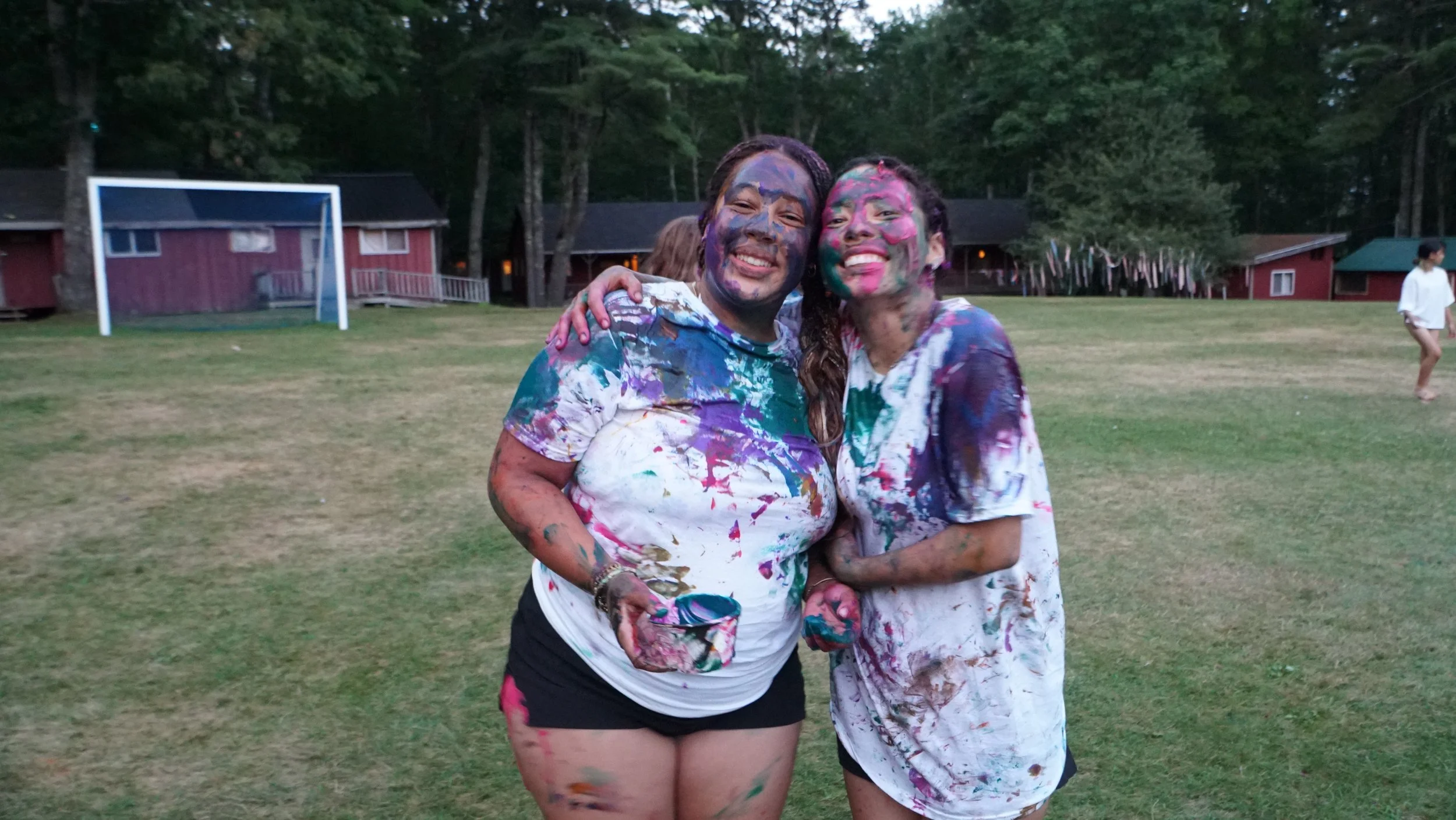 Two women covered in colorful paint, smiling and hugging at an outdoor campfire area with small buildings and a soccer goal in the background.