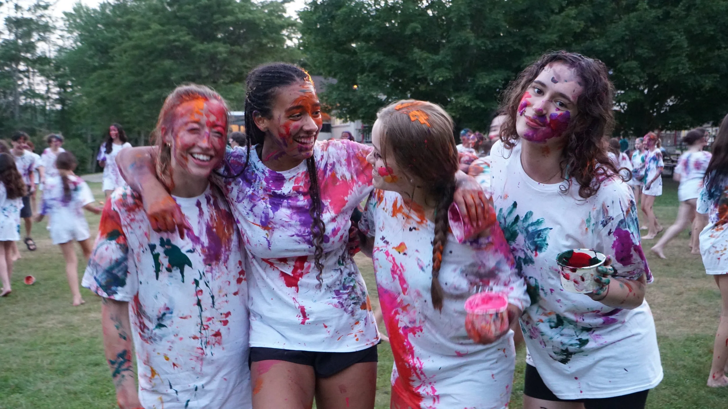Group of four smiling girls with paint on their faces and clothes, celebrating outdoors on grass with trees in the background, during a colorful paint throw event.