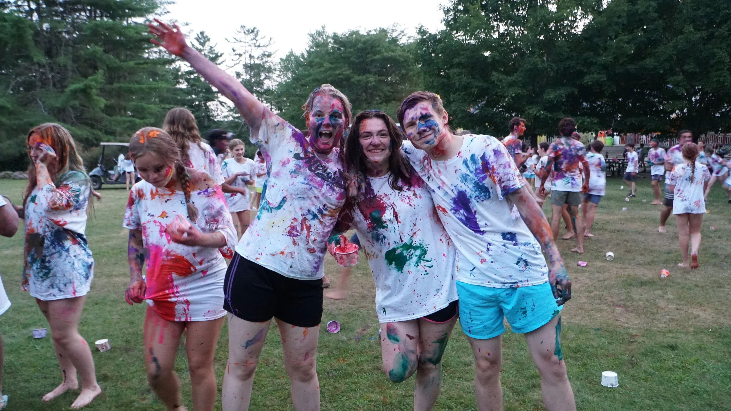 People covered in colorful paint at an outdoor event on a grassy field, celebrating and smiling.