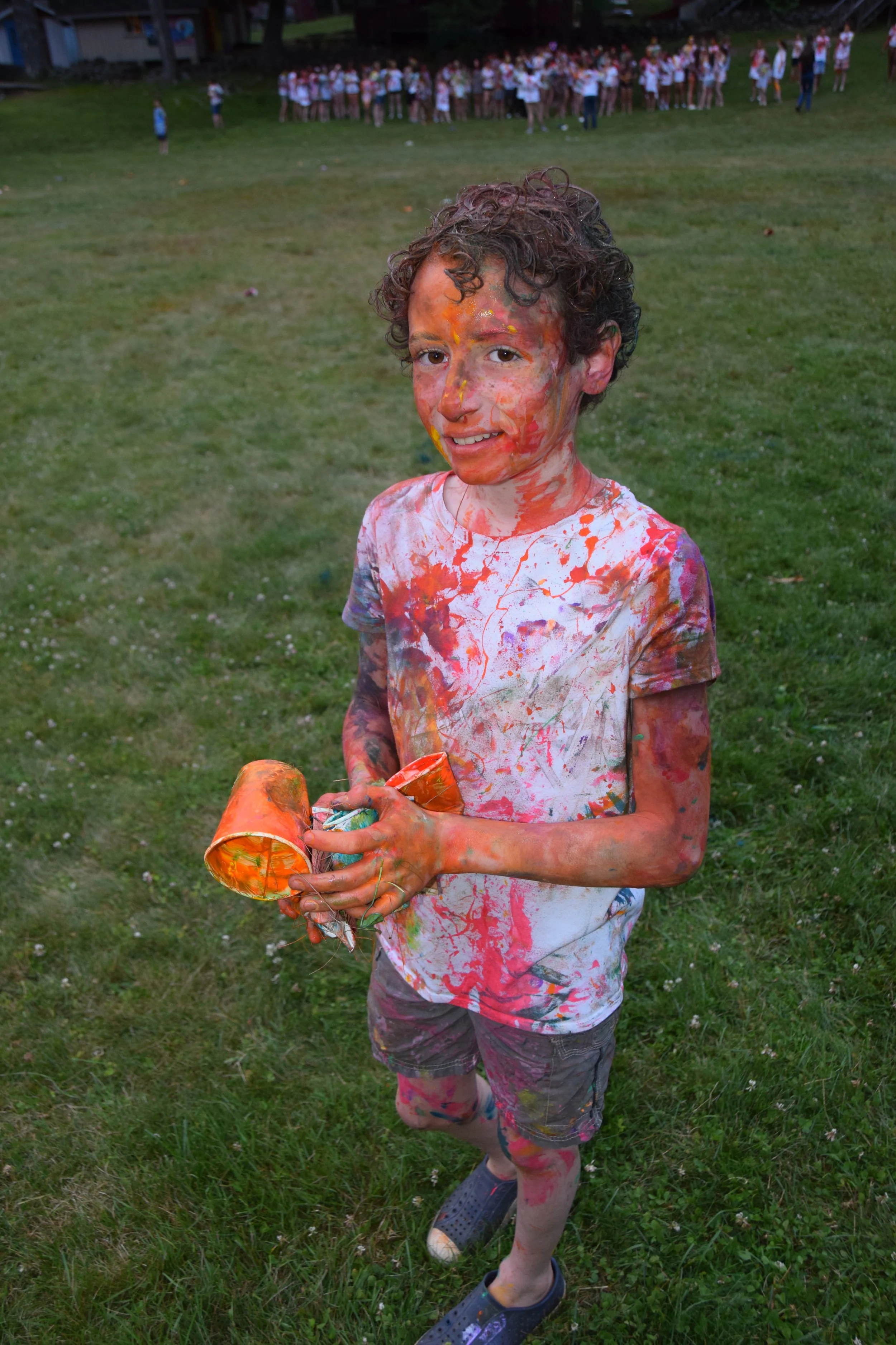 A young boy covered in colorful paint, holding a hat, standing on a grassy field during an outdoor event with a group of children in the background.