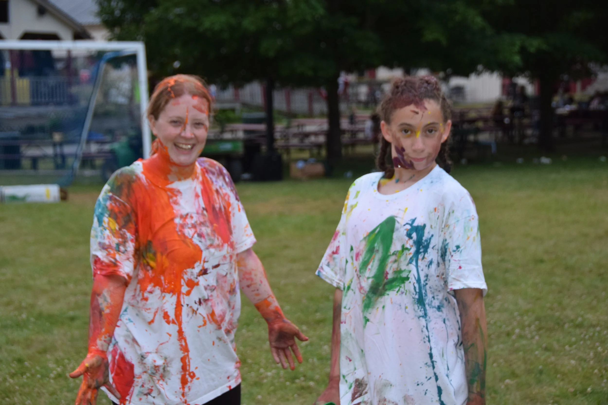 Two women with paint-covered clothes and faces standing on a grassy field, smiling and posing for the camera.