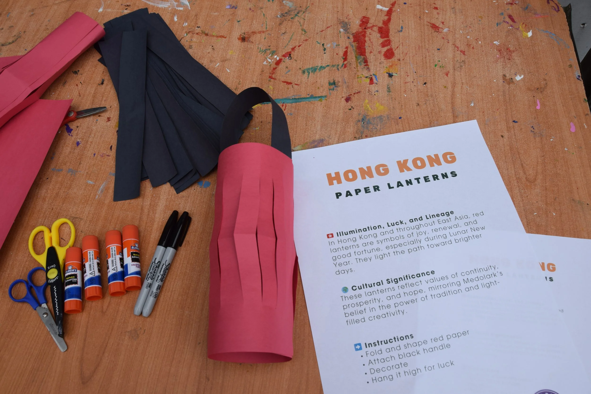 Materials for making paper lanterns on a wooden table, including pink and navy paper, scissors, glue sticks, markers, and instructions for crafting a "Hong Kong Paper Lantern."